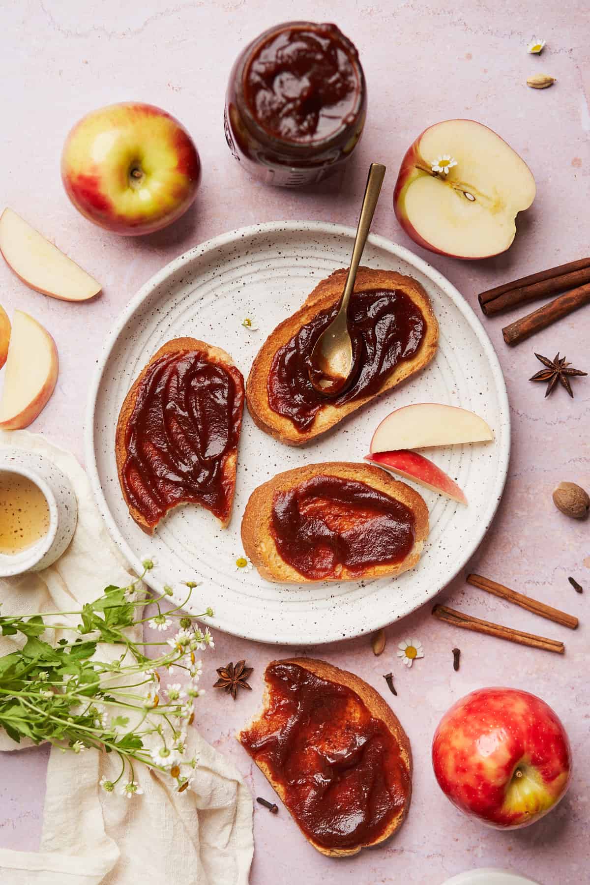 pieces of toast topped with freshly made apple butter, surrounded by fresh apples, cinnamon sticks, cloves, star niase, and maple syrup.