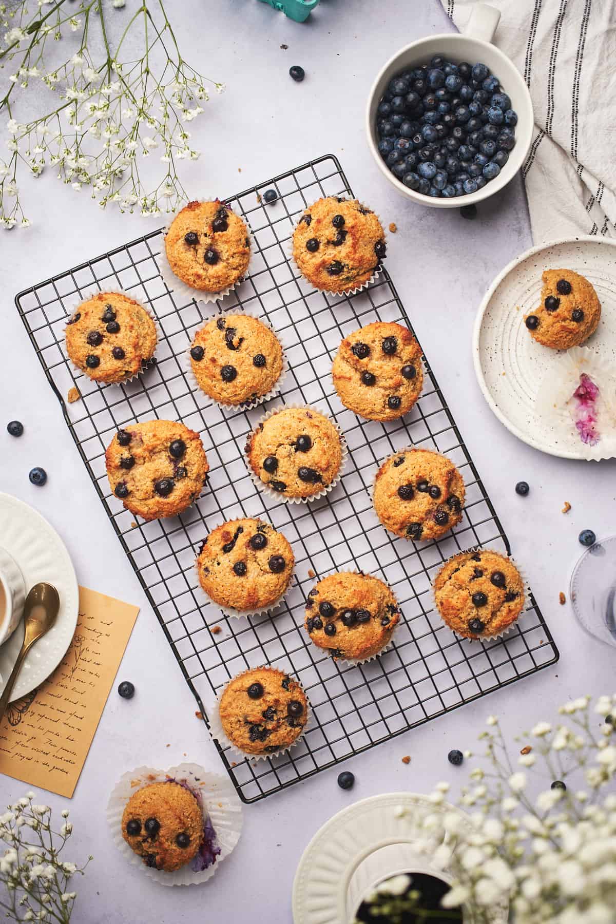 keto blueberry muffins on a wire cooling rack with a plate nearby with a half eaten muffin, fresh blueberries, white flowers, and coffee.