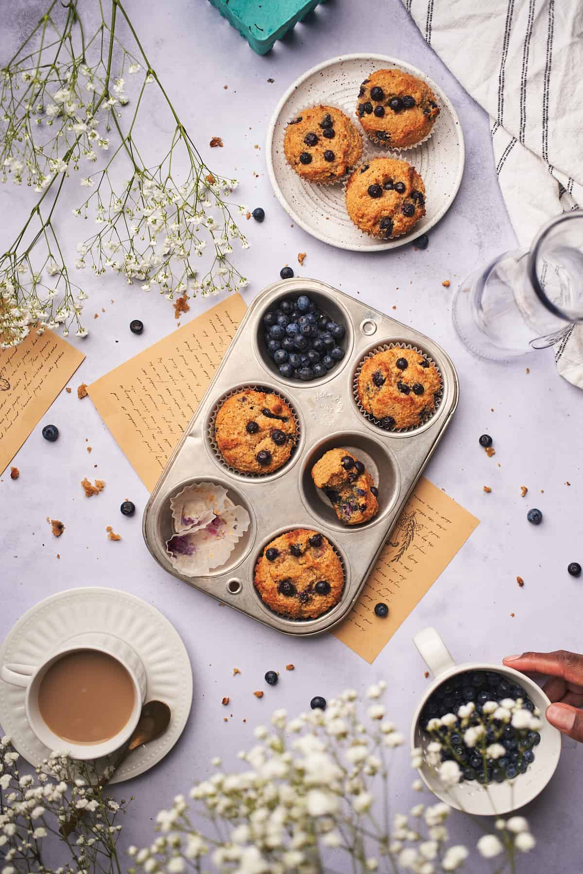 hand reaching a coffee cup overlooking a lovely scene of keto blueberry muffins with coffee and flowers. 
