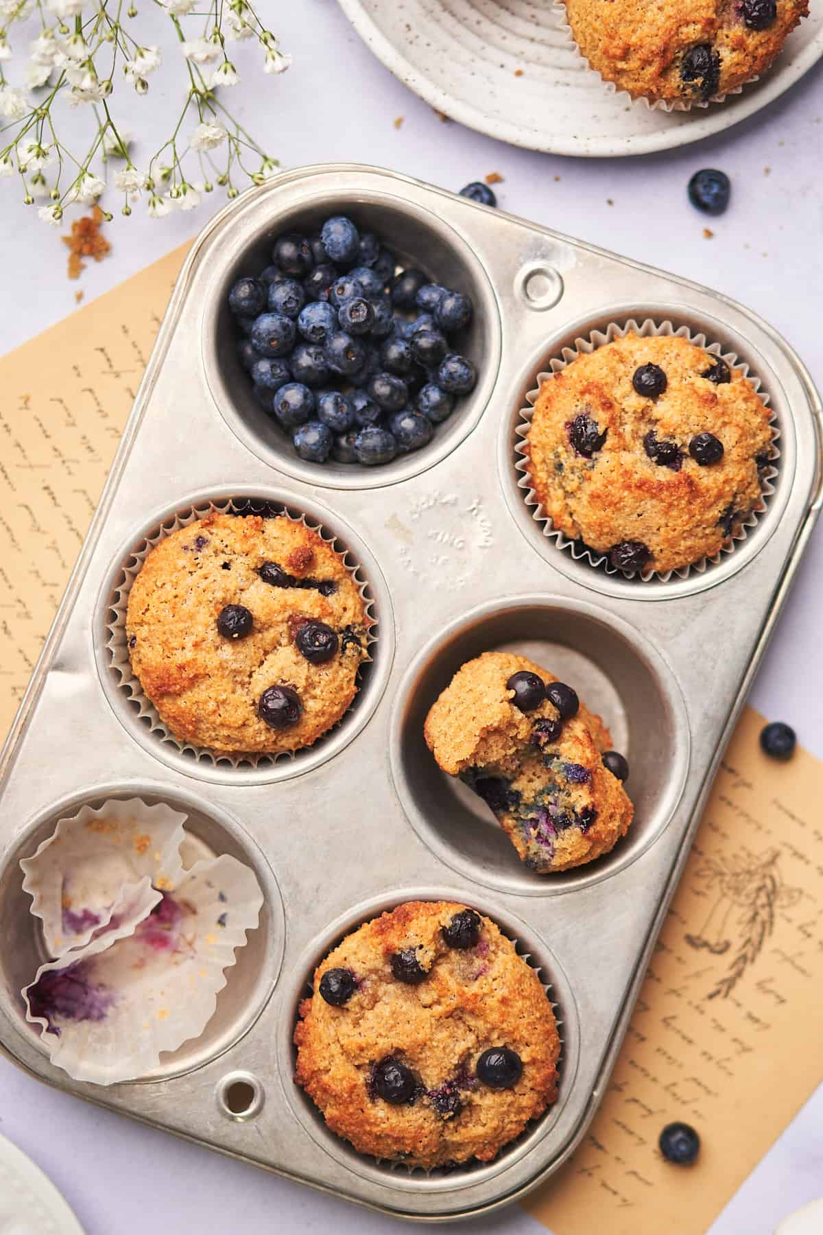 close up shot of keto blueberry muffins in a muffin tin with fresh blueberries, and one of the muffins with a bite taken out of the center. 