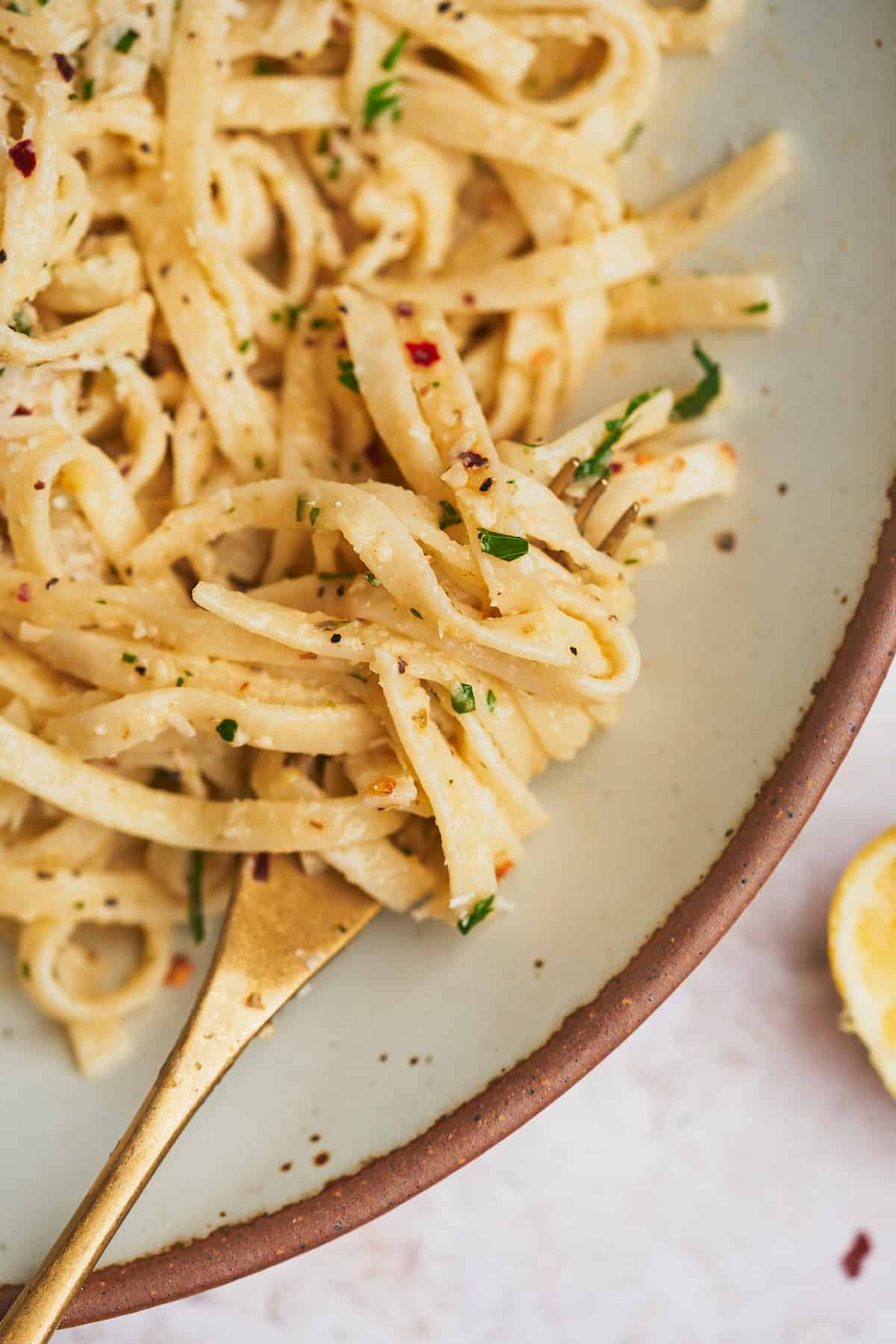 close up macro shot of a plate of linguine pasta topped with parmesan, parsley, and red pepper flakes wrapped around a gold fork. 