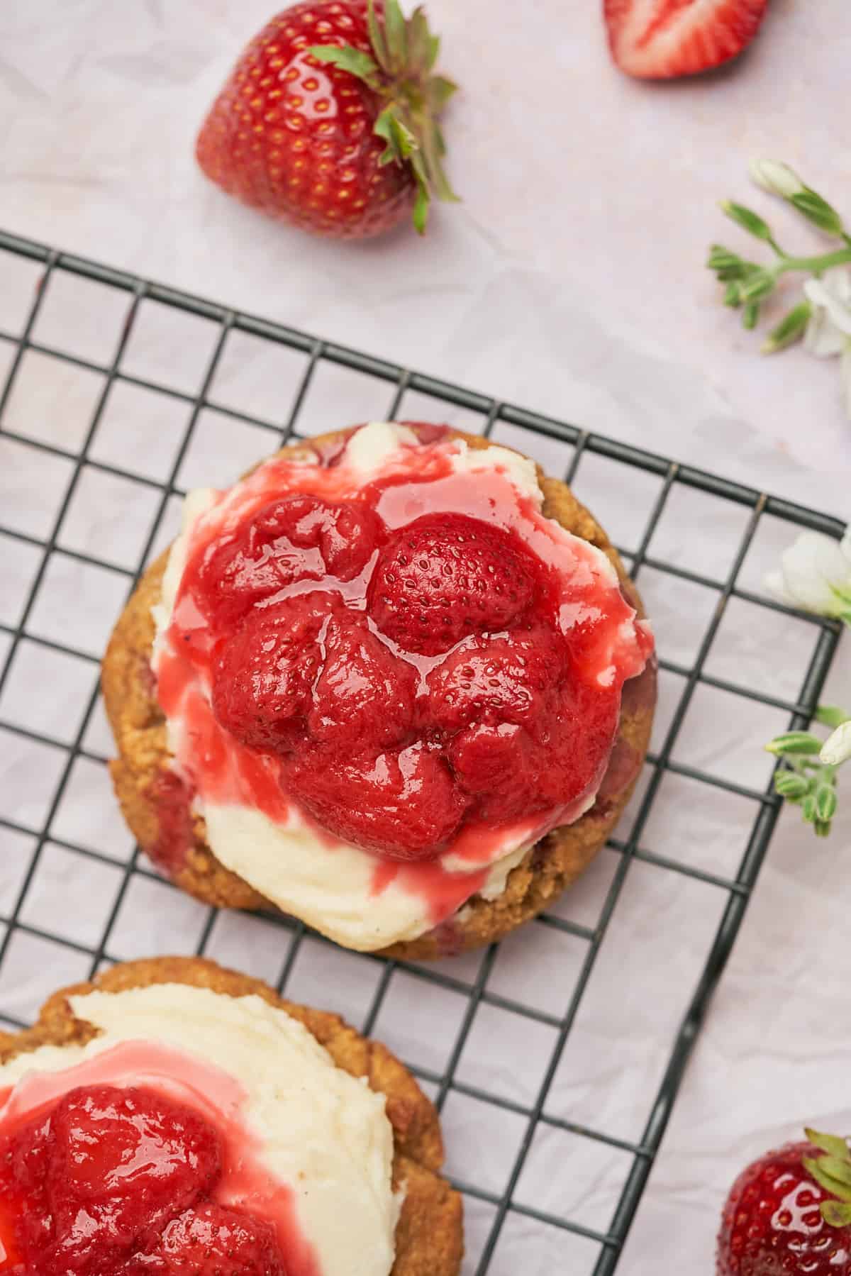 gorgeous strawberry cheesecake cookies with fresh strawberry topping drizzling off the sides on a cooling rack, surrounded by white flowers. 