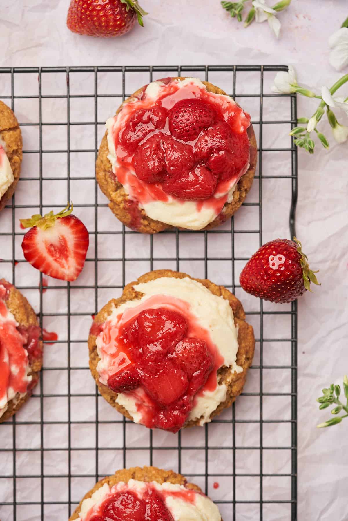 strawberry cheesecake cookies with fresh strawberry sauce, strawberries cut in half with a cream cheese frosting, on a black metal cooling rack, and white flowers surrounding. 