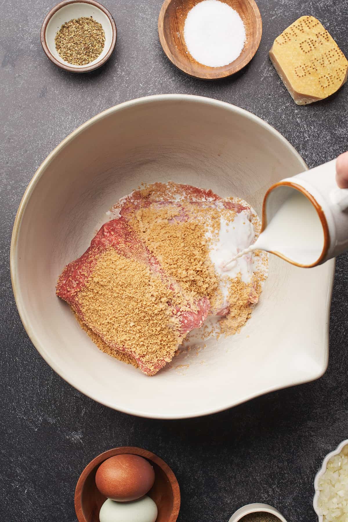 hand pouring milk into a large bowl with raw meat, and breadcrumbs.