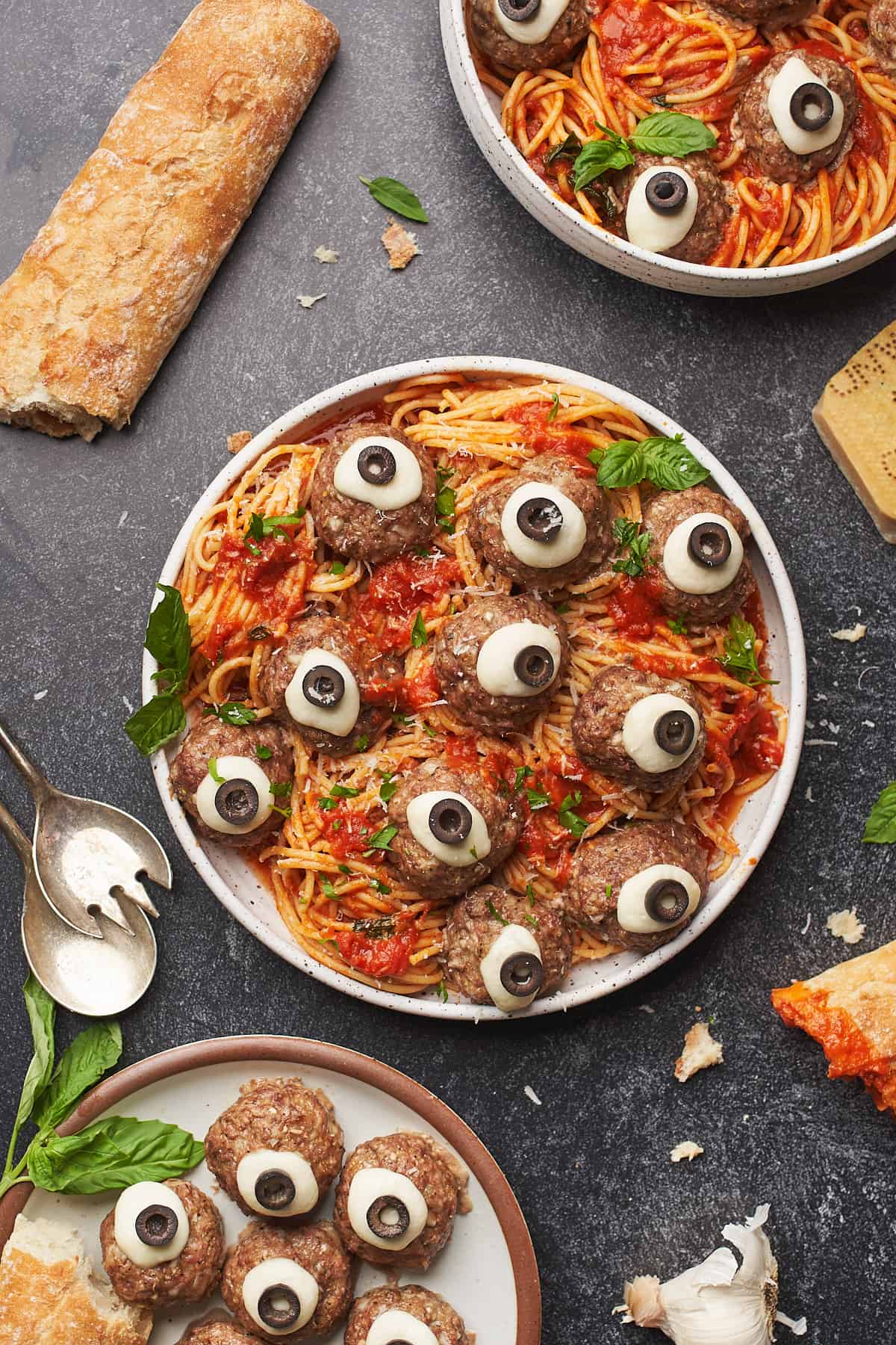 plate full of spaghetti and meatball eyeballs, with mozzarella cheese and olives, surrounded by bread, basil and parmesan cheese. 