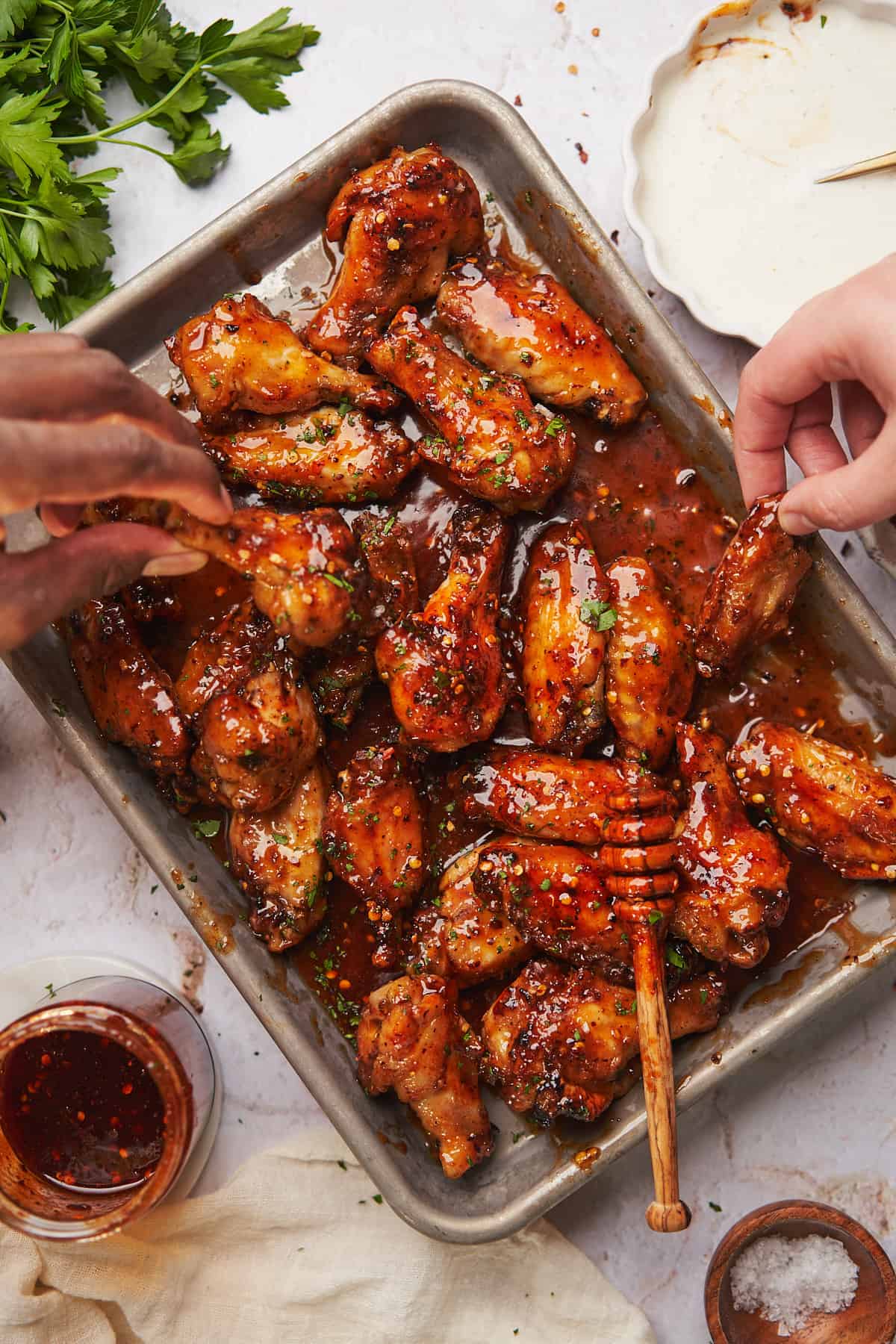 two hands grabbing for sticky hot honey chicken wings laid out on a baking sheet. 