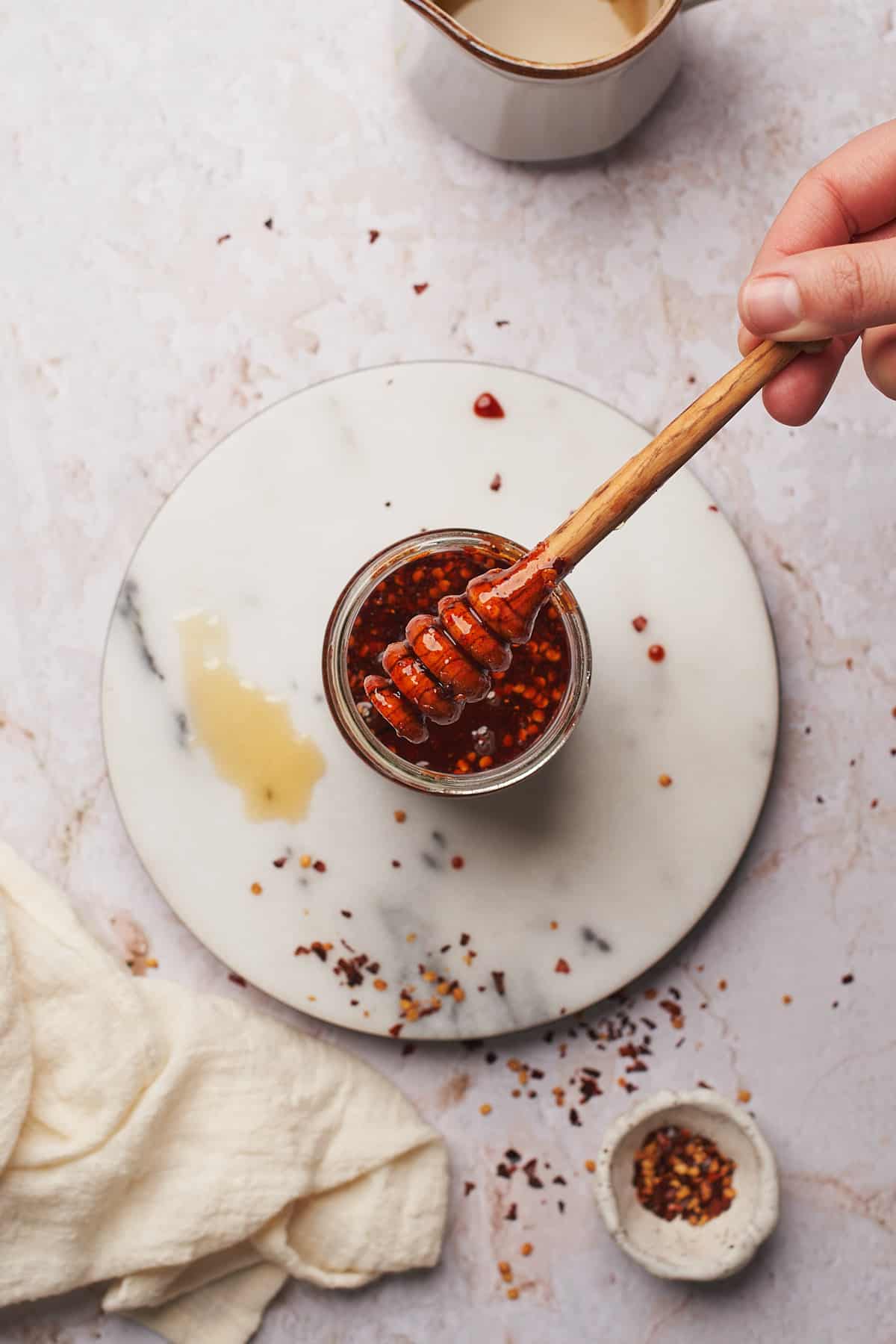 hand holding a wooden honey dripper in a small glass jar with red pepper flakes around it. 