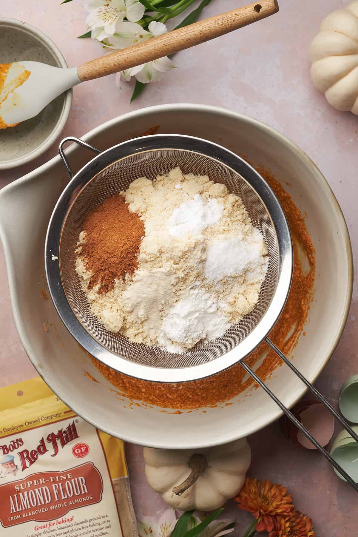 dry ingredients in a metal sieve over a large mixing bowl with a bag of bob's red mill almond flour nearby. 