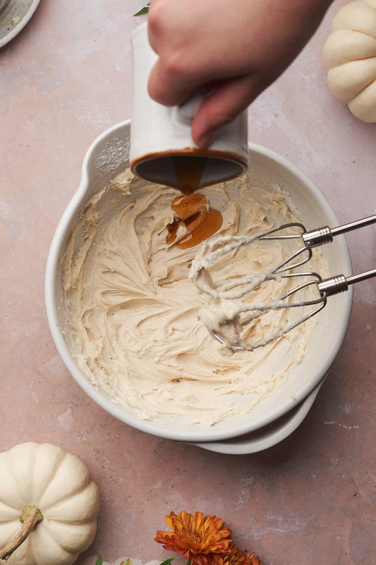 hand pouring in maple syrup into a bowl of cream cheese frosting. 