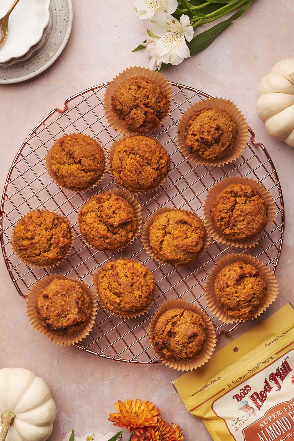 pumpkin muffins cooling on a round rose gold cooling rack, surrounded by a bag of Bob's red mill almond flour, and a white pumpkin. 