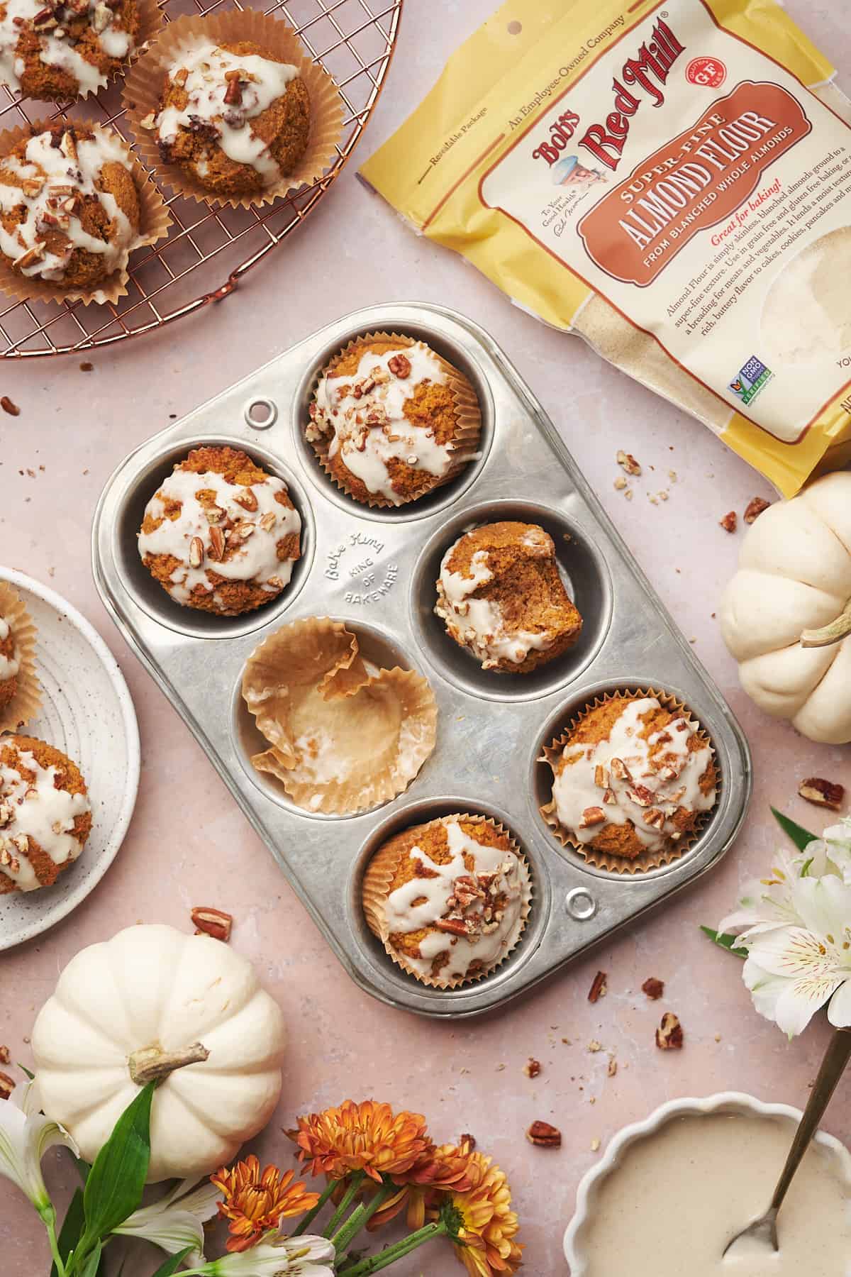 flat lay shot of keto pumpkin muffins topped with a cream cheese glaze and pecans, surrounded by a bag of bob's red mill almond flour, flowers, and white pumpkins. 