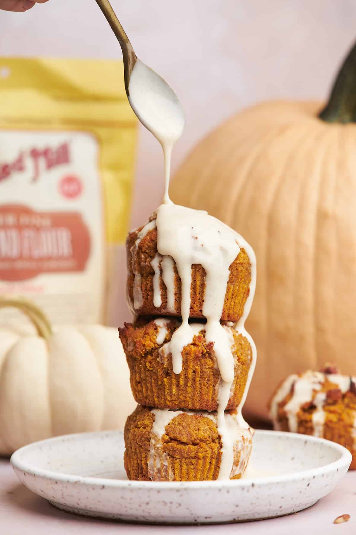 bag of bob's red mill almond flour in the background of a stack of pumpkin muffins being drizzled with cream cheese glaze. Large pumpkins are in the background.