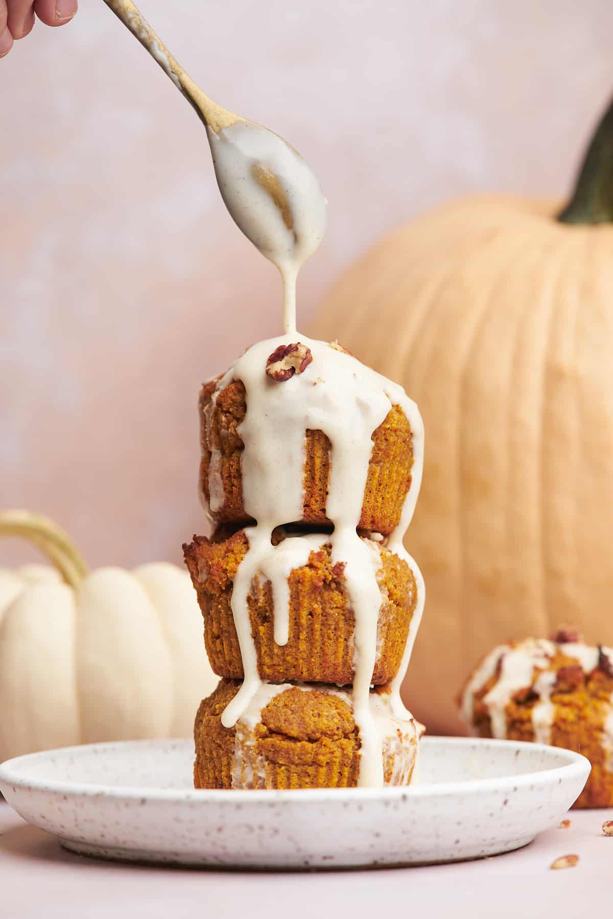 stack of pumpkin muffins with a hand holding a small spoon over top drizzling cream cheese glaze over the stack, and pumpkins in the background. 