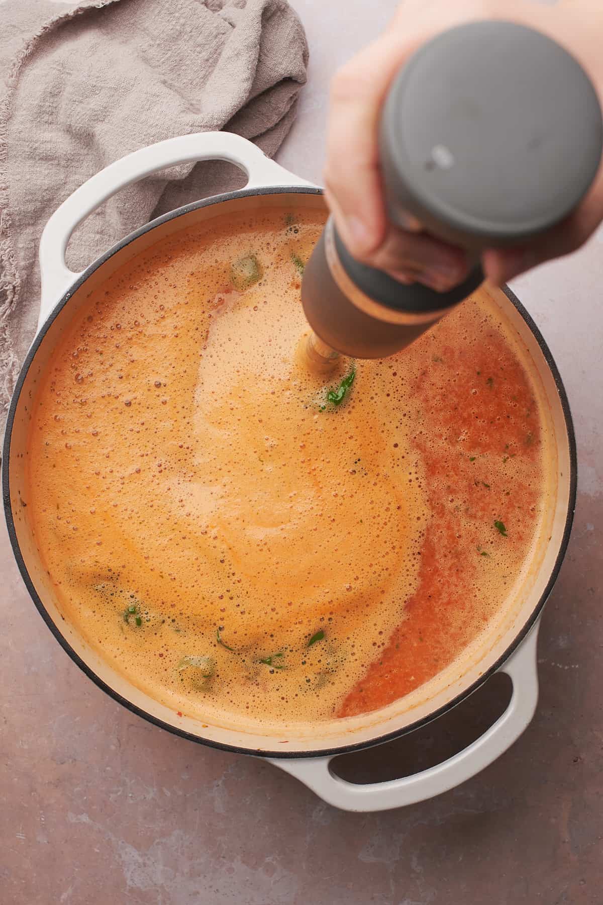 hand holding an immersion blender and pureeing tomato soup in a pot. 