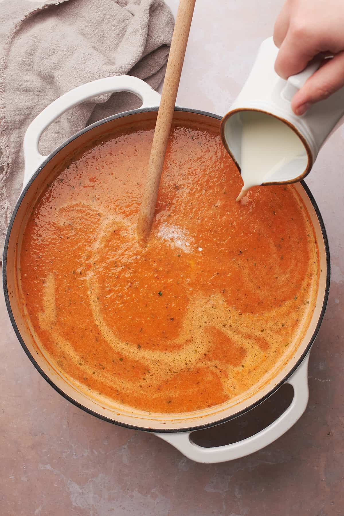 hand pouring heavy cream into a dutch oven full of tomato basil soup. 