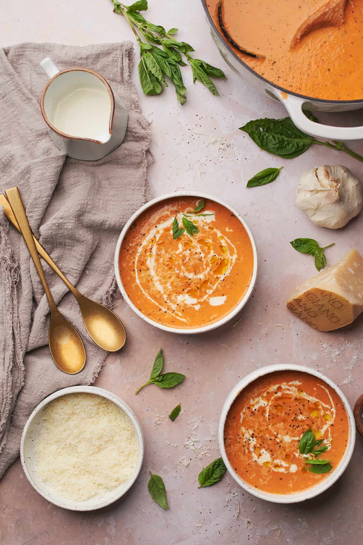 bowls of creamy tomato soup with cream, olive oil, basil, parmesan, and garlic surrounding it.