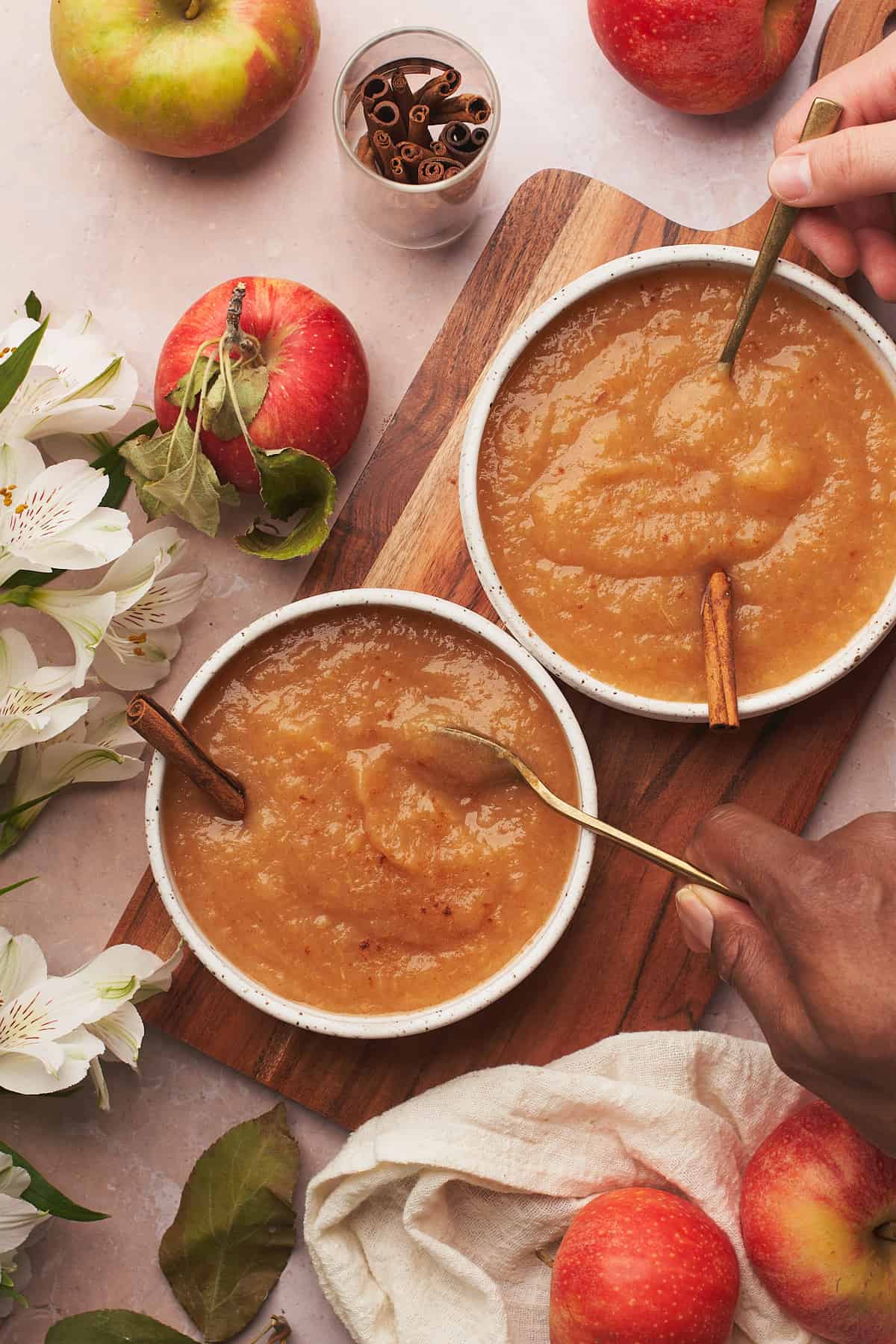 two people digging into bowls of applesauce, surrounded by white flowers and fresh apples. 