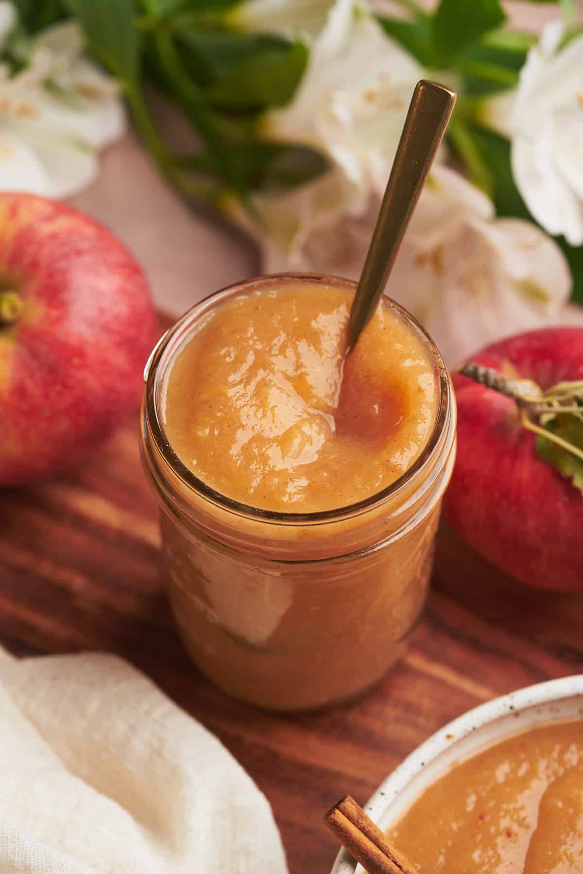 fresh apple sauce in a mason jar surrounded by apples, white flowers, and a bowl of applesauce. 