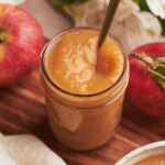 fresh applesauce in a mason jar, surrounded by apples, white flowers, and a nice wooden board.