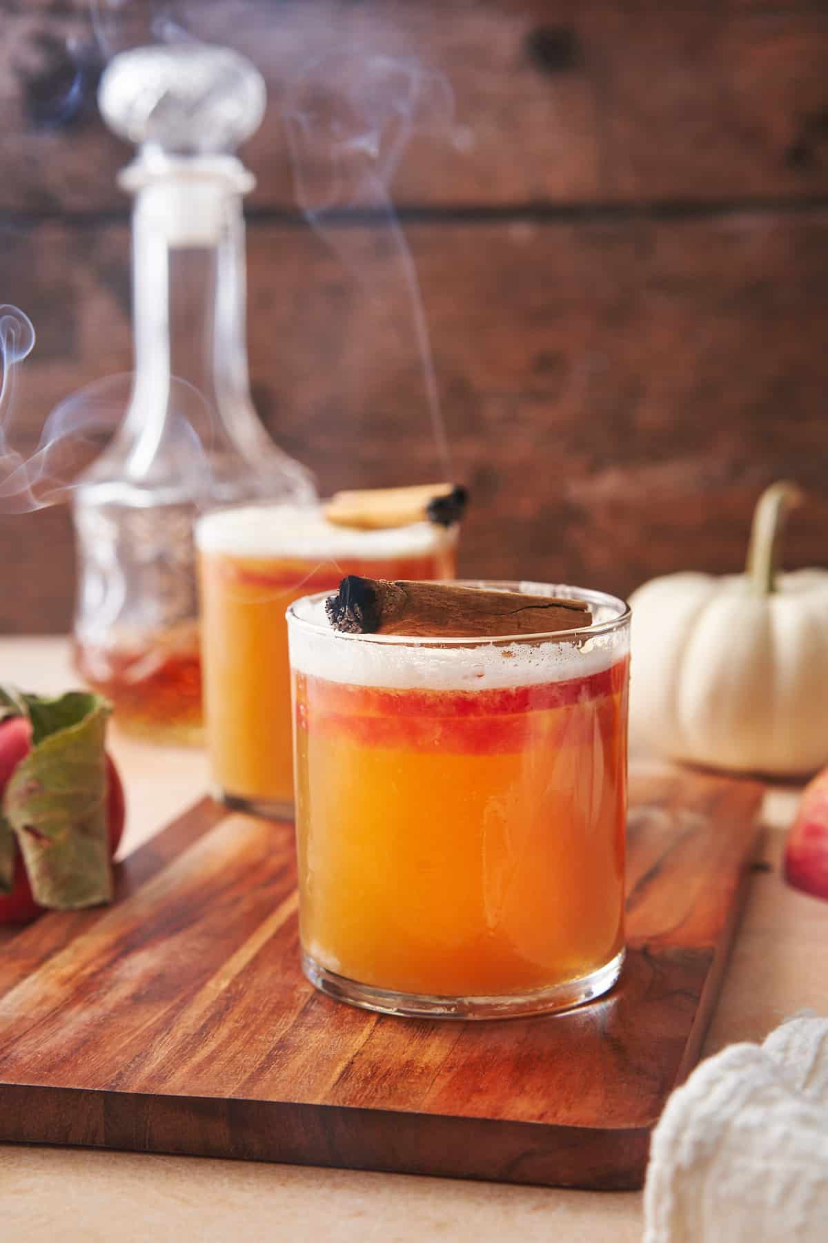 apple whiskey sour with a gorgeous fall backdrop, a white pumpkin, whiskey, and apples in the background, topped with a smoking cinnamon stick. 
