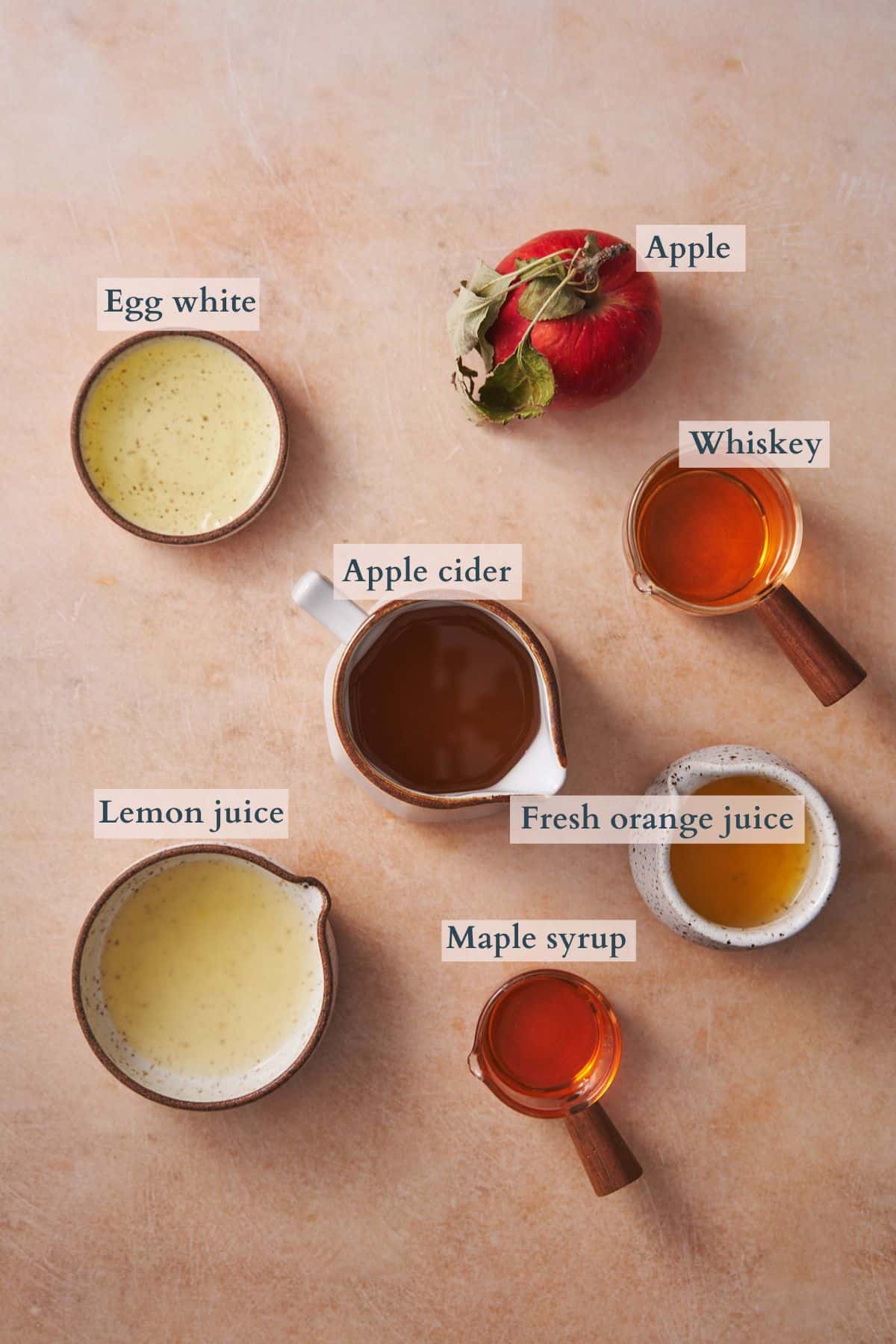 ingredients to make an apple whiskey sour laid out on a table in ceramic dishes labeled with text to denote the different ingredients. 