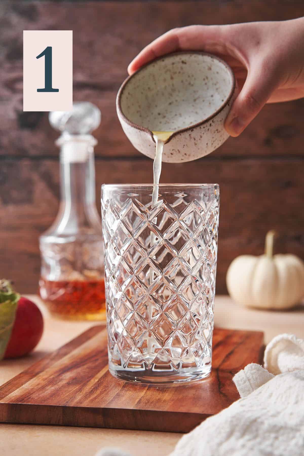 hand pouring lemon juice into a cocktail shaker with a lovely scene with pumpkins, whiskey, dark wood, and apples in the background.