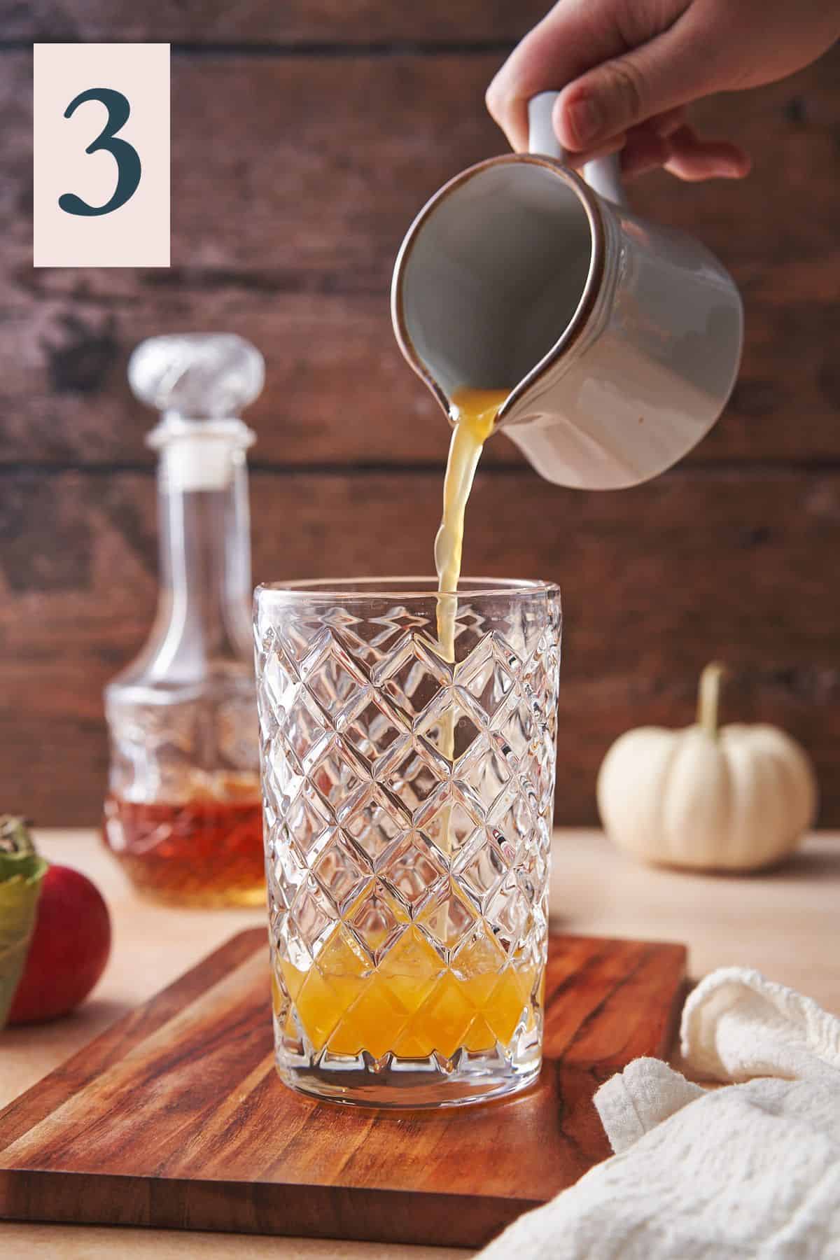 hand pouring apple cider from a ceramic pourer into a cocktail shaker with a lovely scene with pumpkins, whiskey, dark wood, and apples in the background.