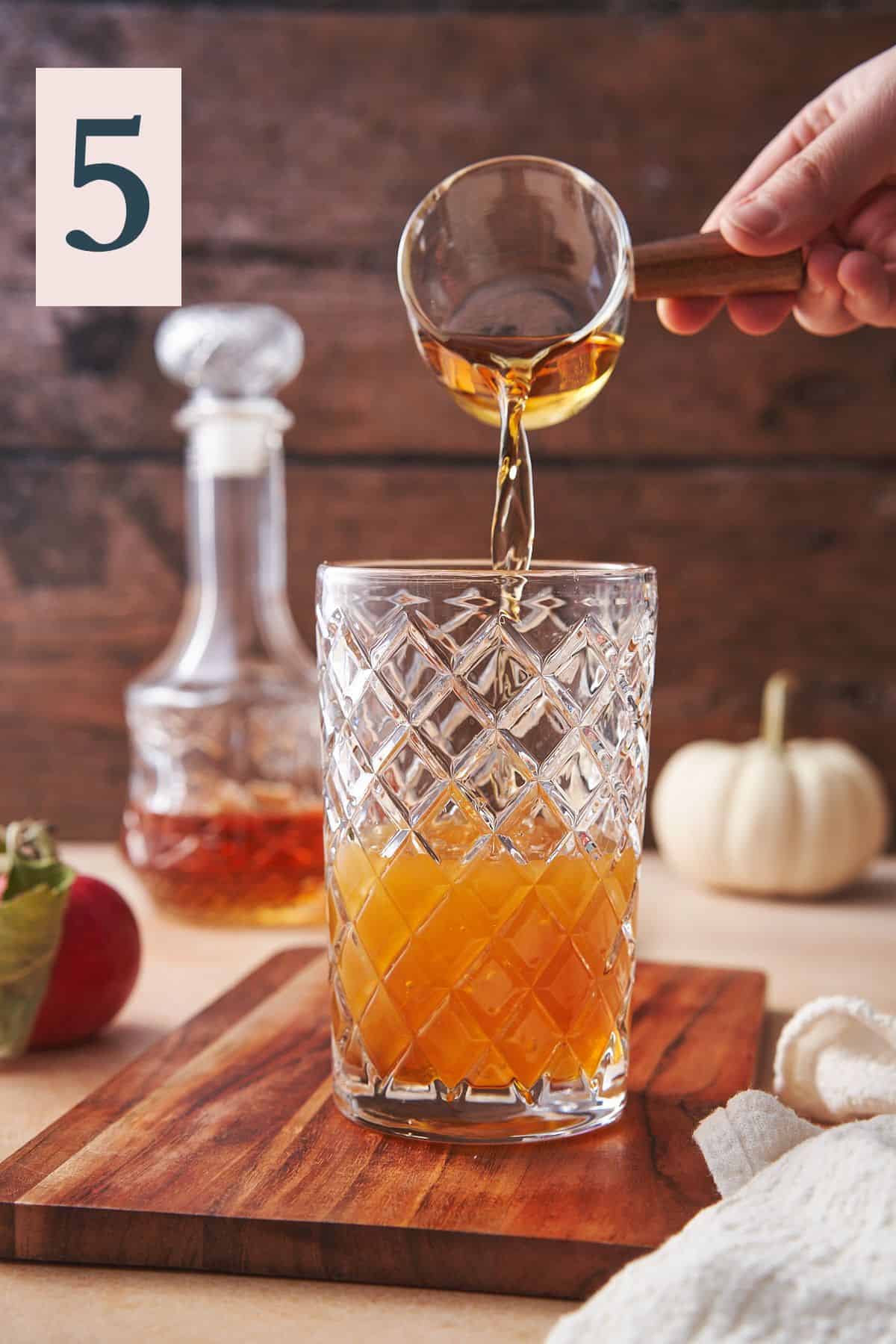 hand pouring bourbon from a glass and wood container into a cocktail shaker with a lovely scene with pumpkins, whiskey, dark wood, and apples in the background.