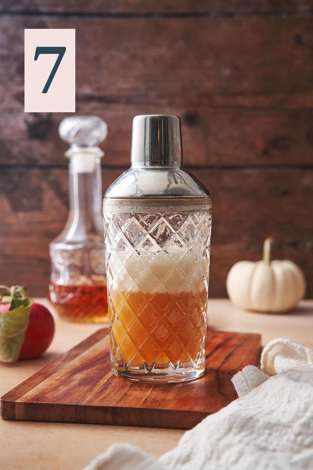 glass cocktail shaker on a wooden board, with a dark wood backdrop, bourbon, a white pumpkin and an apple with a foamy top on the mixture.