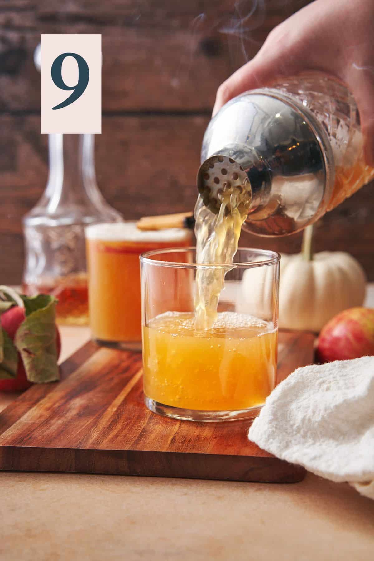 pouring an apple whiskey sour into a glass with one made in the background, and a nice scene with pumpkins, whiskey, and apples.