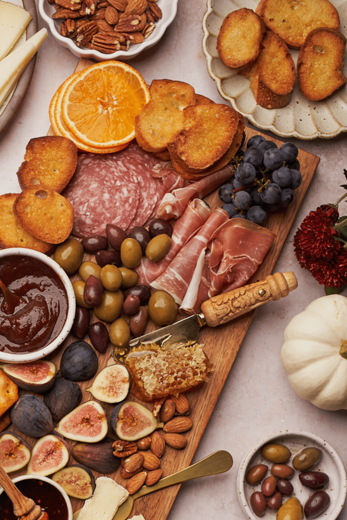fall charcuterie board with a small white pumpkin, red mums, meats, cheeses, olives, grapes, crackers, honeycomb, and more. 