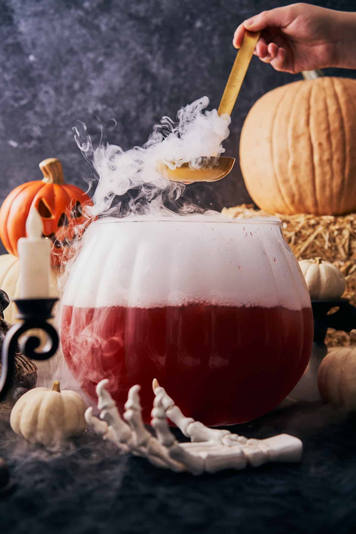 spooky halloween punch inside of a pumpkin shaped punch bowl with dry ice coming out, and pumpkins in the background. 