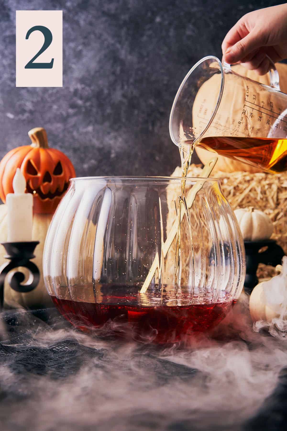 hand pouring spiced rum into a large pumpkin shaped punch bowl, with a creepy jackolantern in the background and a number 2 in the top left hand corner.