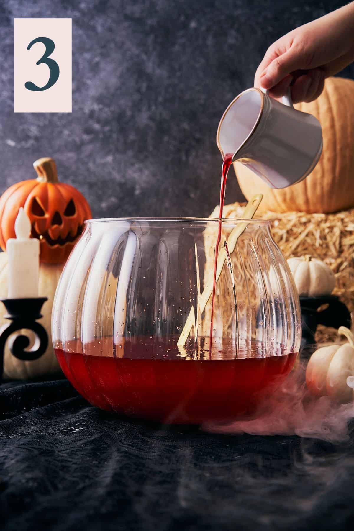 hand pouring grenadine into a large pumpkin shaped punch bowl with a spooky Halloween scene with dry ice and a number 3 in the upper left hand corner.