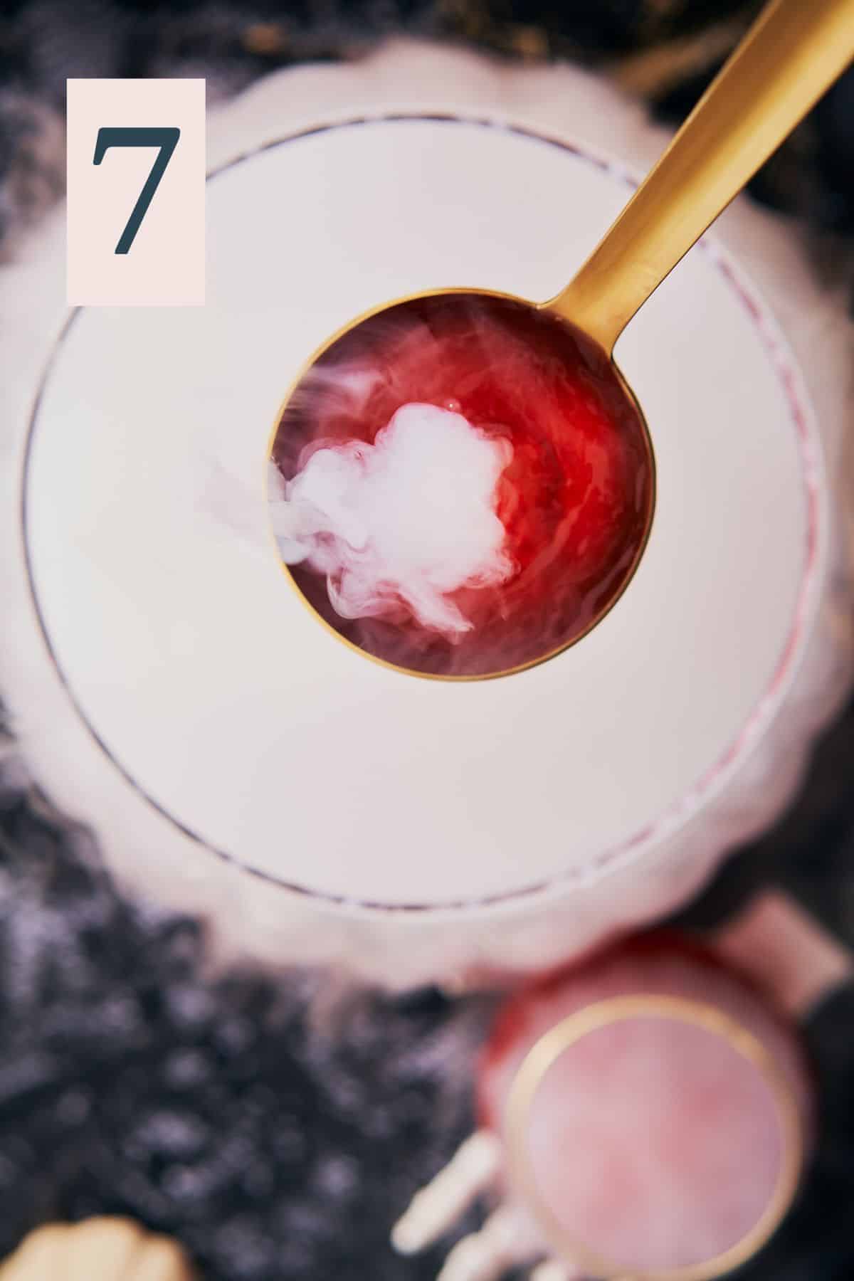 ladle of bright red liquid over a punch bowl with smoking dry ice and a number 7 in the upper left hand corner. 