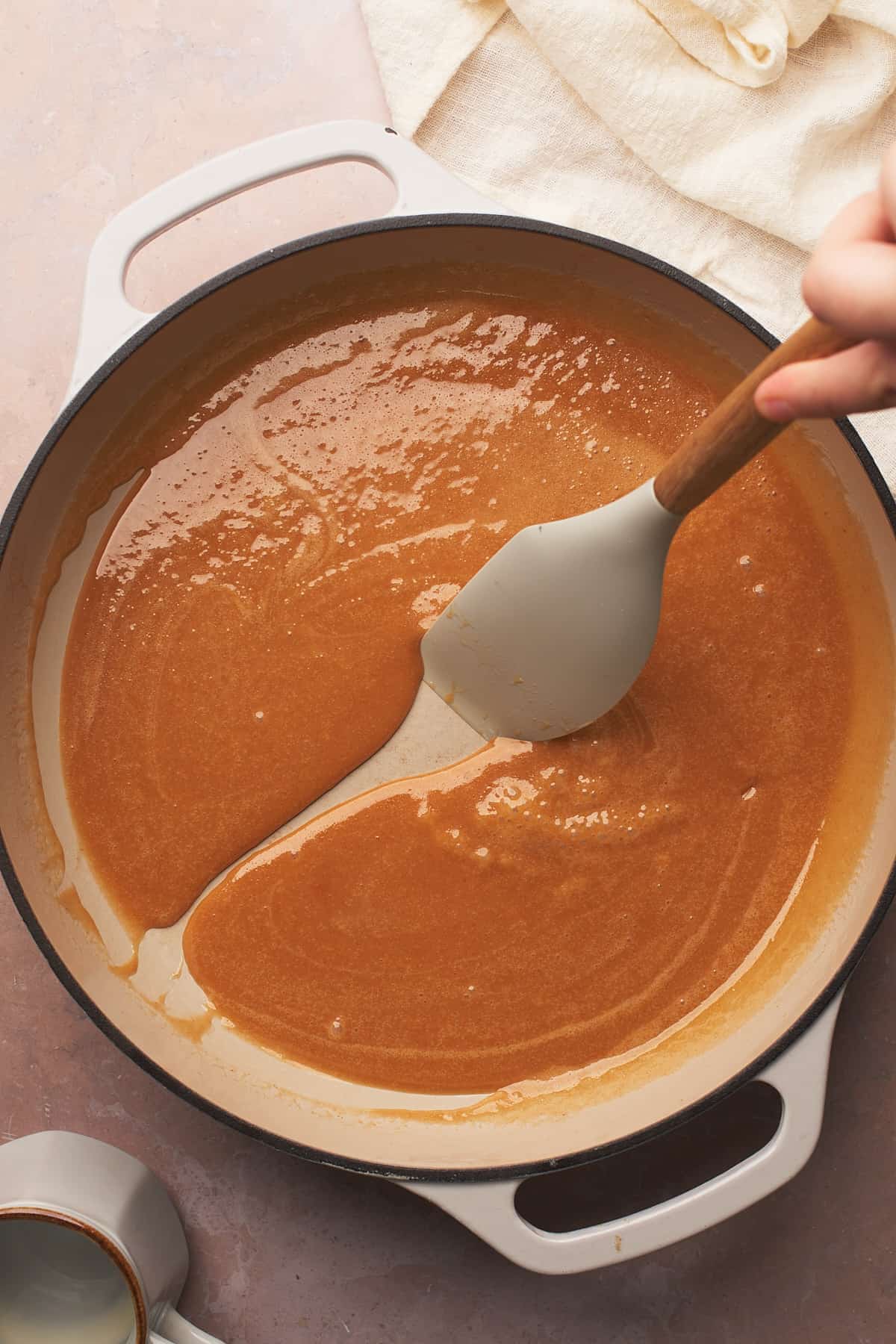 hand dragging a rubber spatula along the bottom of an enameled cast iron skillet to show the thickness of the caramel sauce.