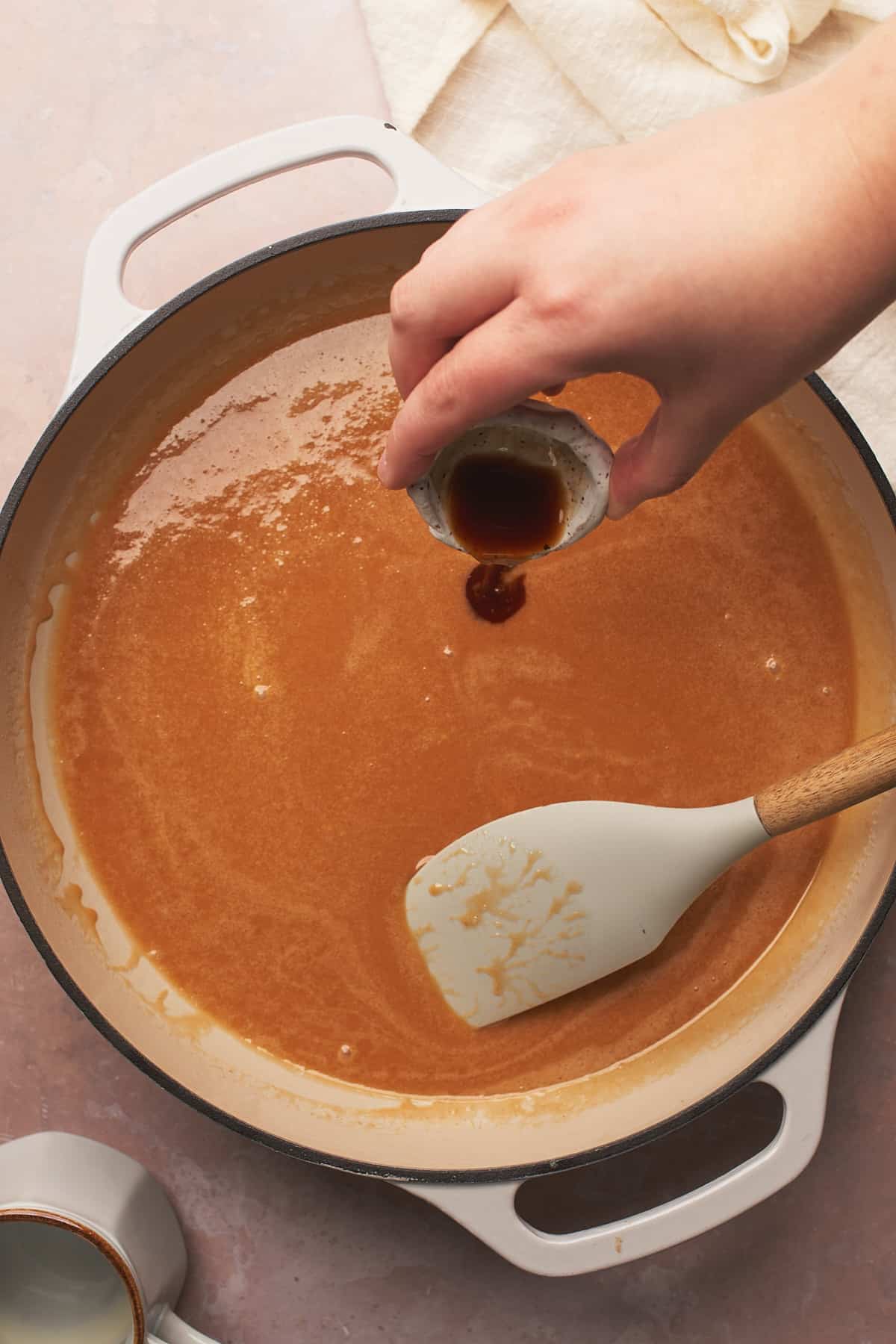 hand pouring vanilla extract into an enameled cast iron skillet full of caramel sauce.