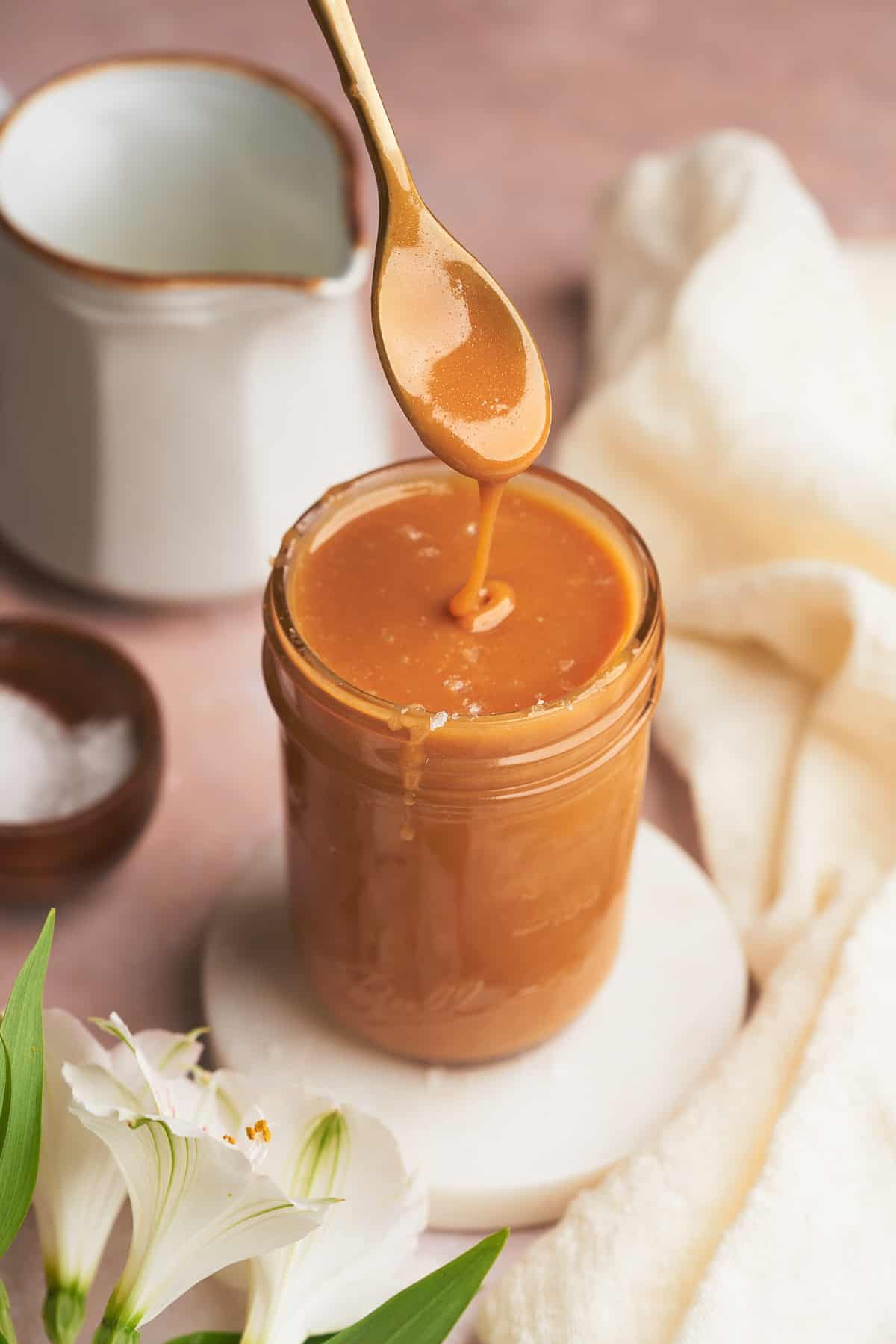 spoon dipping into a jar of salted keto caramel sauce, with with flowers in the foreground and a cream pitcher in the background. 