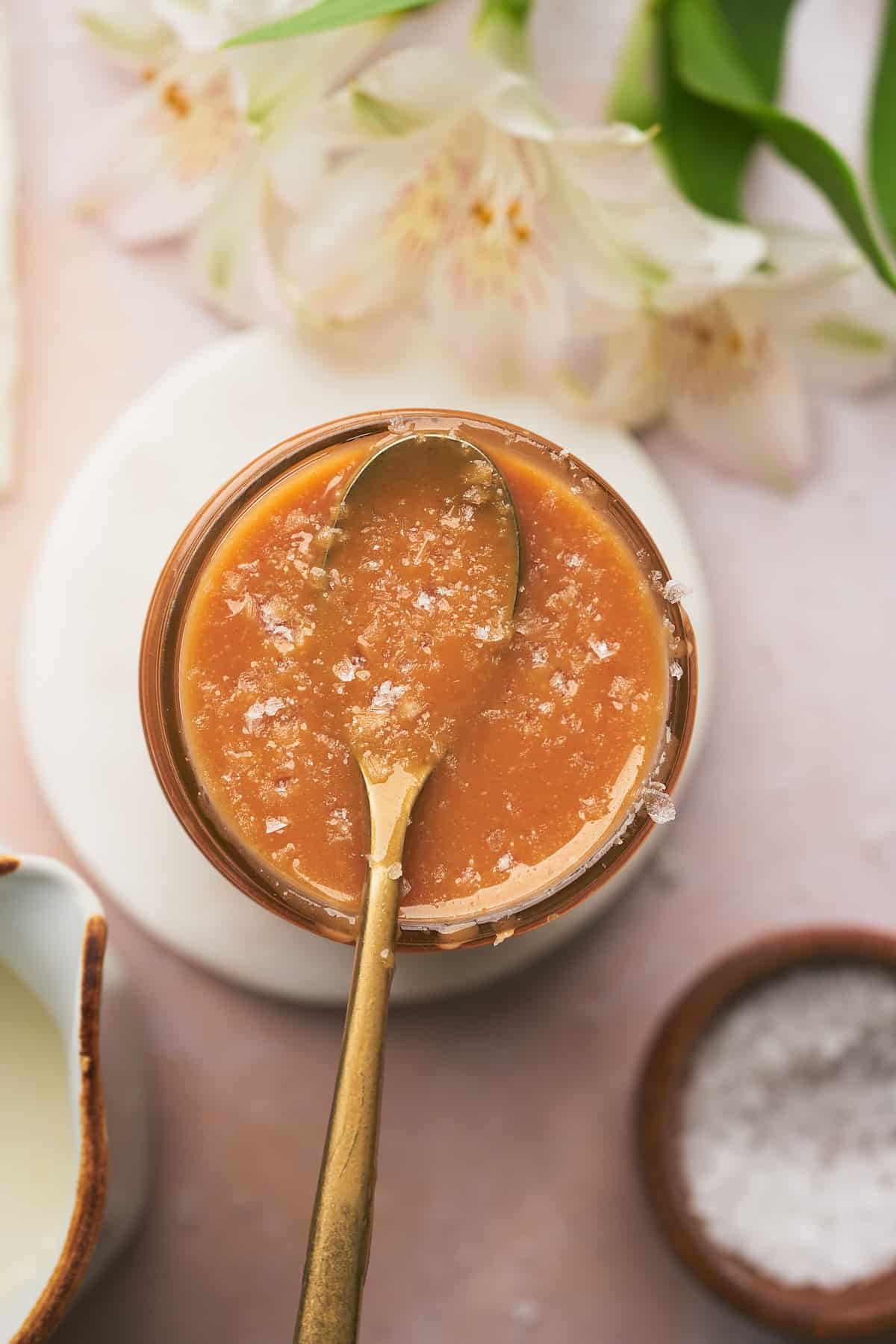 flat lay shot of keto salted caramel sauce in a jar topped with flakey sea salt with white flowers surrounding it. 