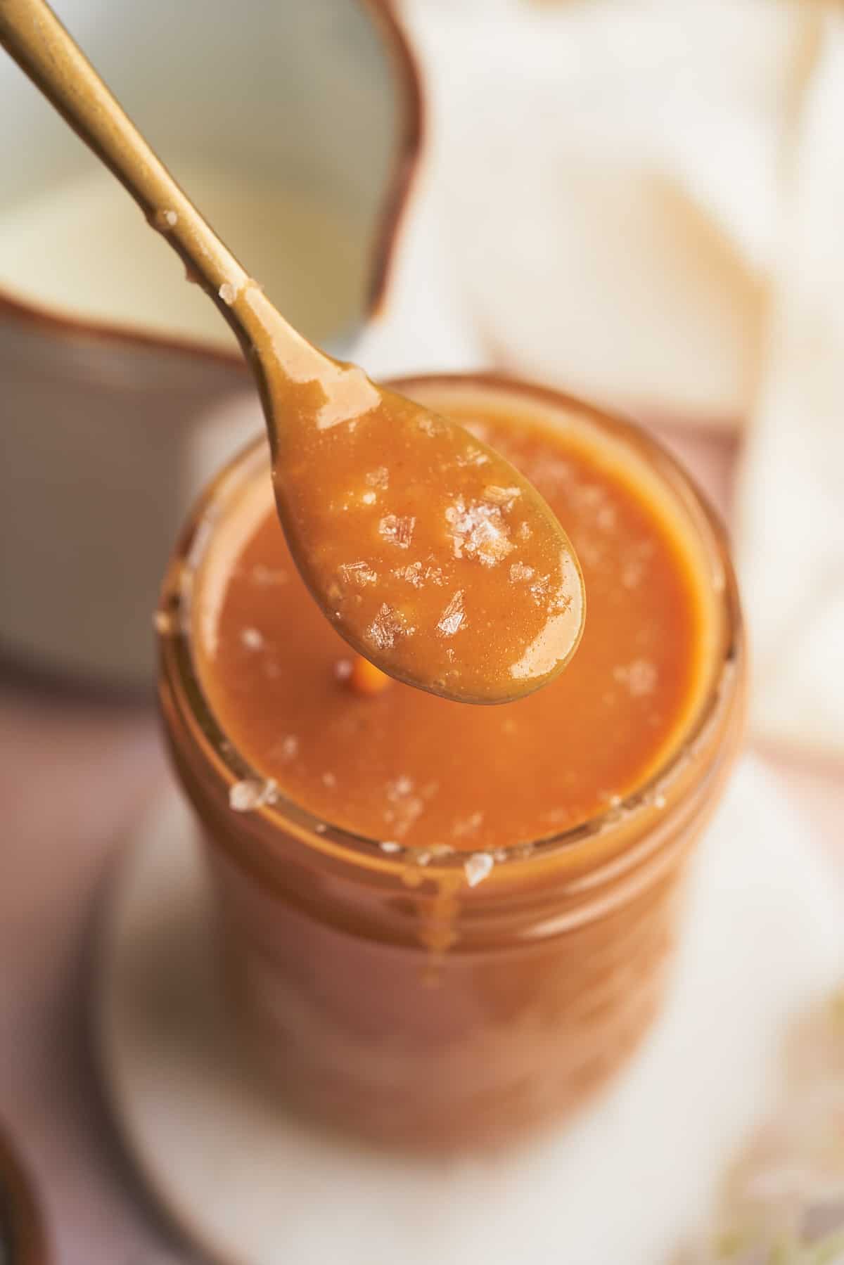 close up shot of a keto salted caramel sauce topped with flaky sea salt on a spoon above a mason jar full of it. 