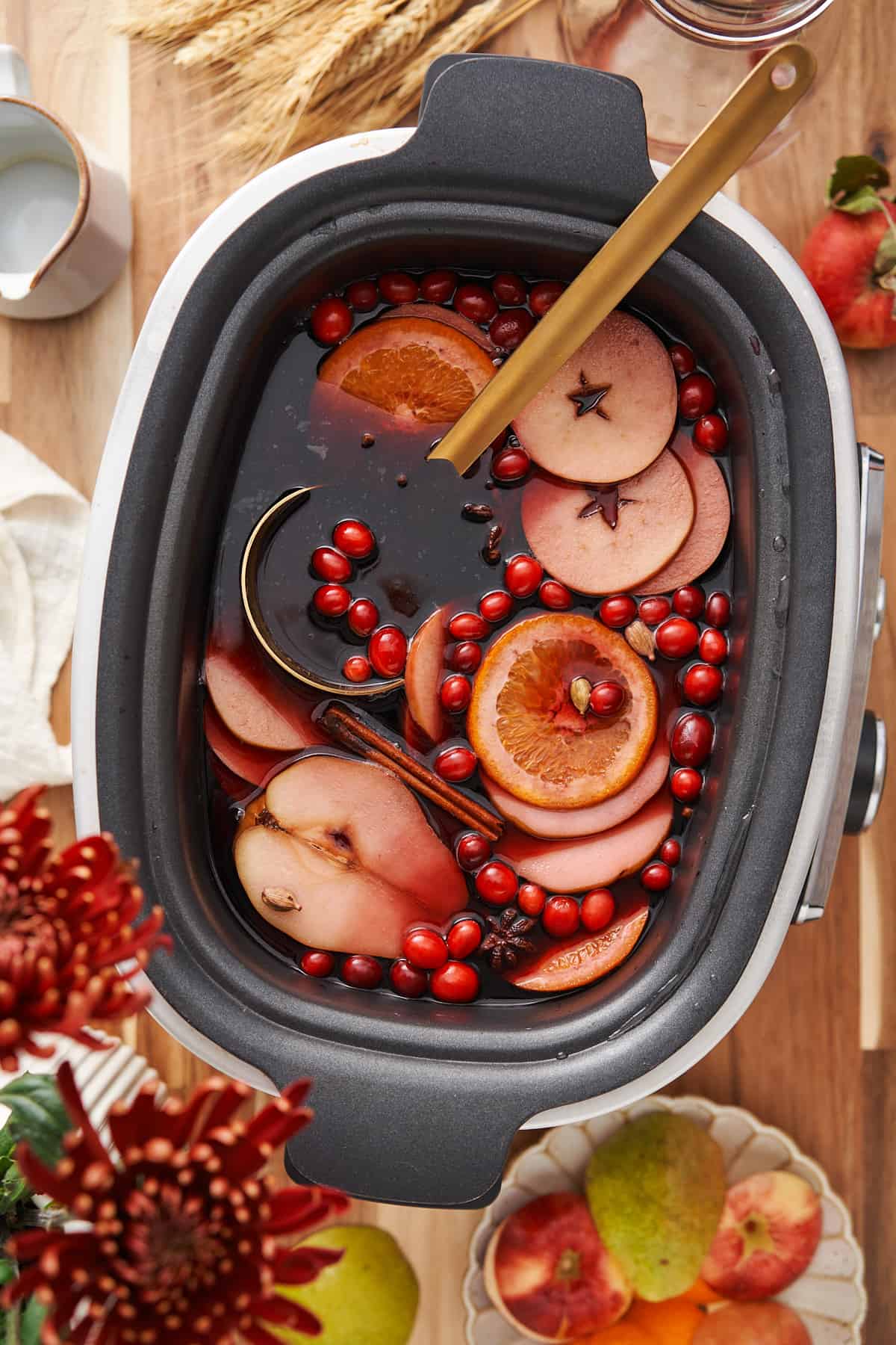 slow cooker mulled wine with a gold ladle inside of the slow cooker, with apple, pear, and orange slices and gresh cranberries.