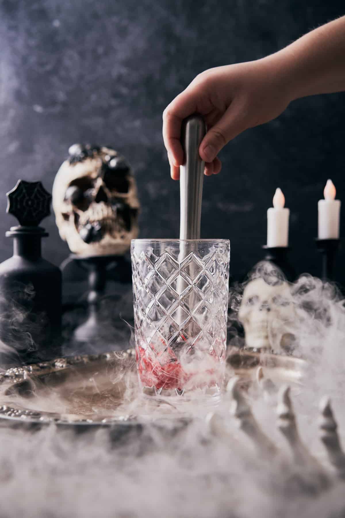 hand muddling raspberries in a glass cocktail shaker, with a very spooky black scene with skulls, candles, and dry ice. 