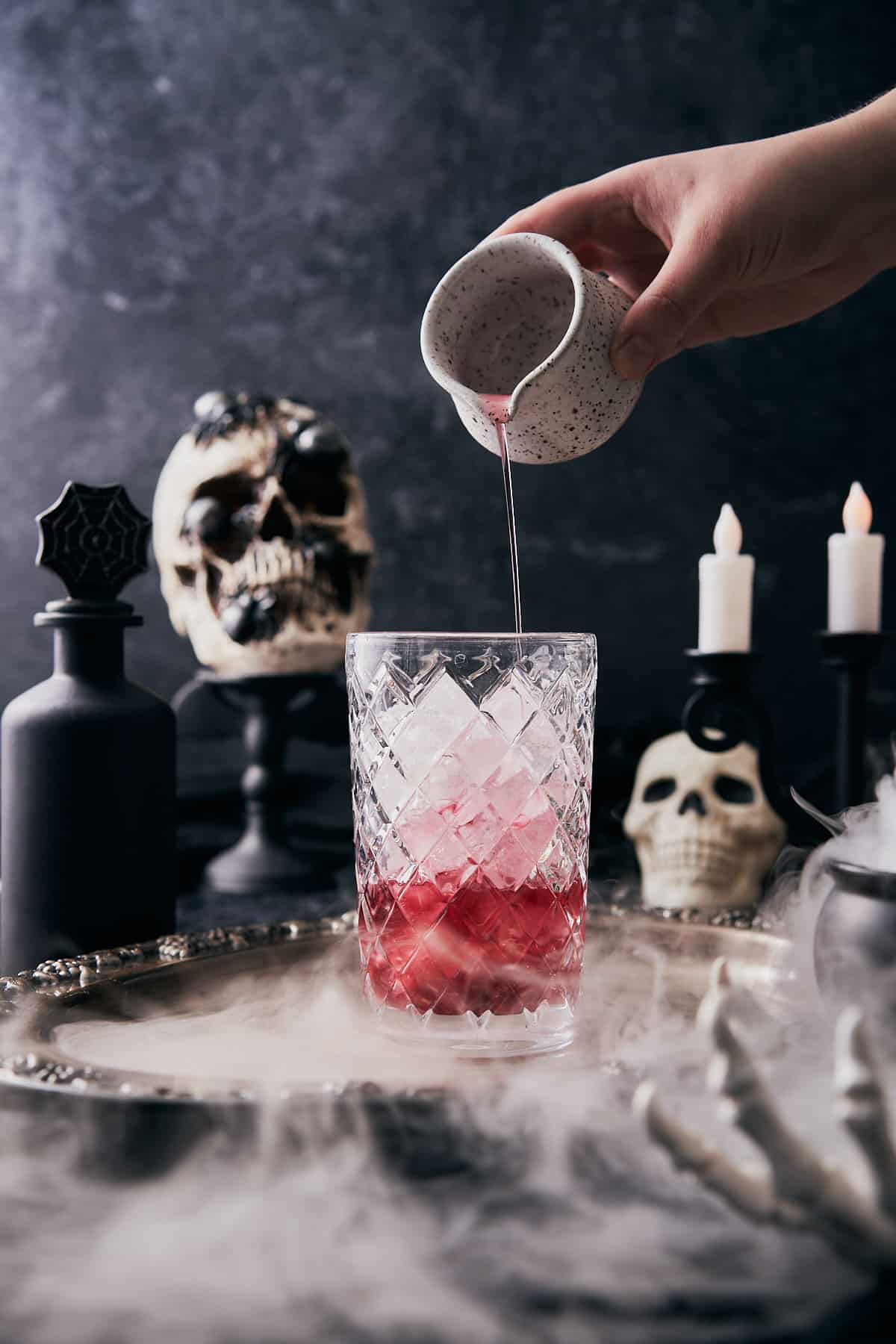 spooky halloween scene with dry ice, and skulls with a hand pouring a red juice into an ice filled cocktail shaker. 
