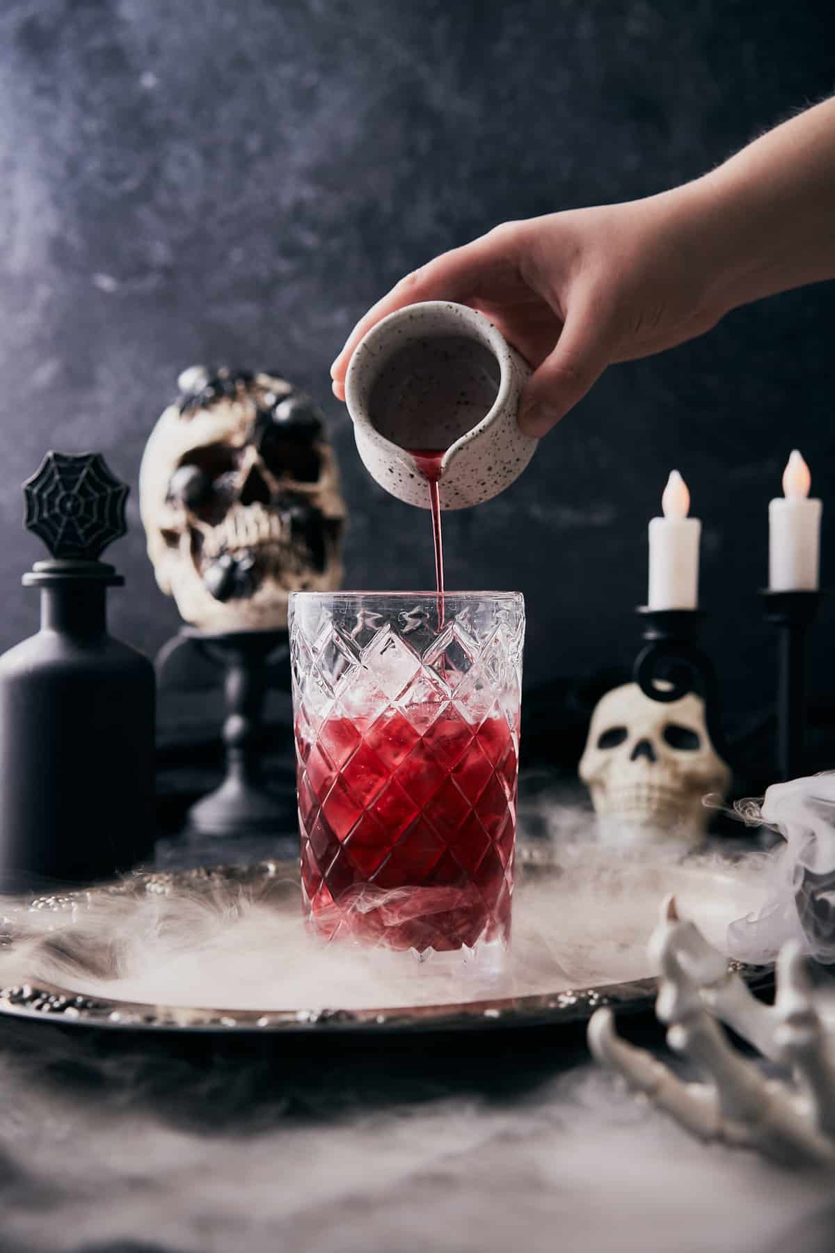 hand pouring grenadine into a glass cocktail shaker with ice in a very spooky scene with dry ice and skulls. 