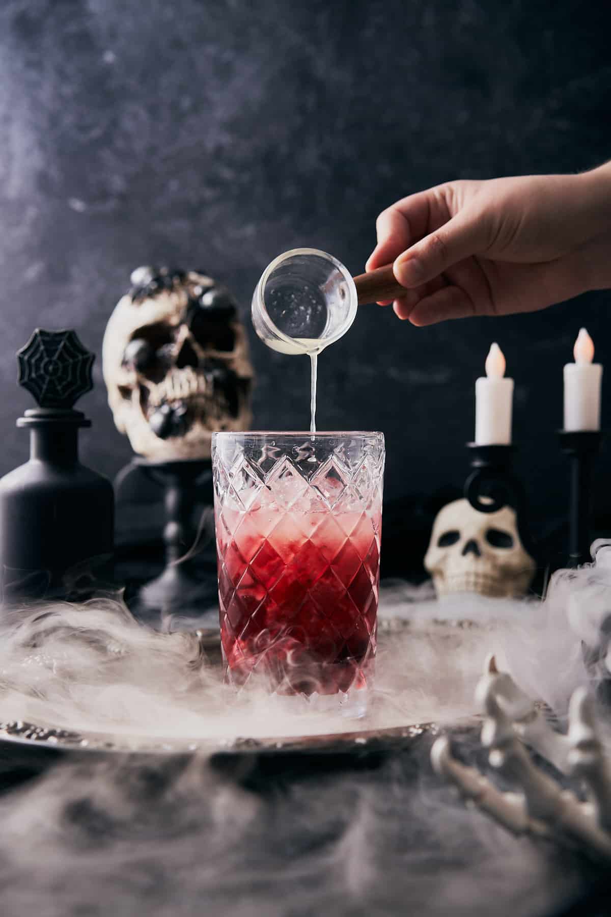 hand pouring lemon juice into a cocktail shaker with a spooky halloween scene in the background. 