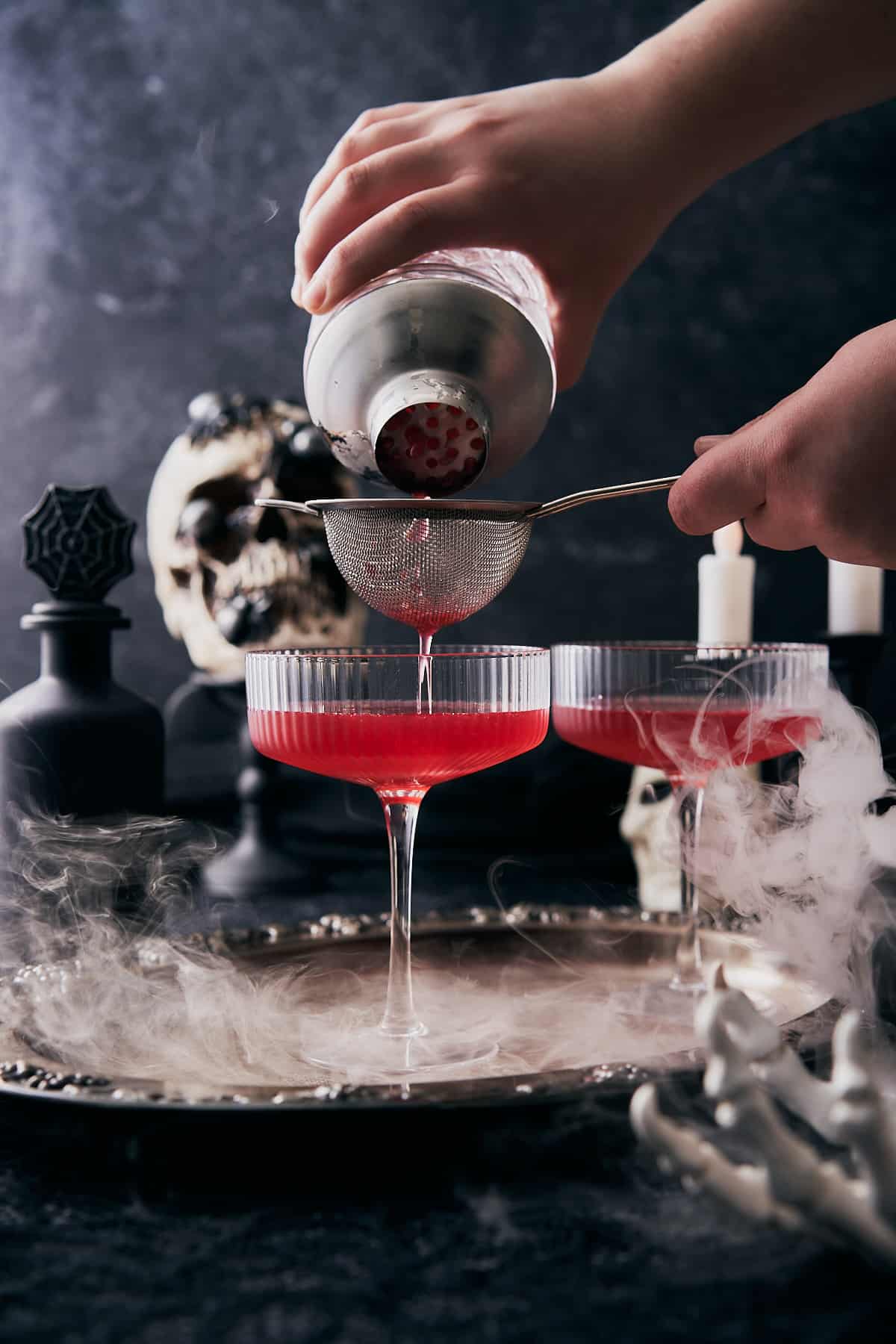 hand double straining a cocktail into a couple glass with a spooky halloween scene surrounding it and dry ice. 