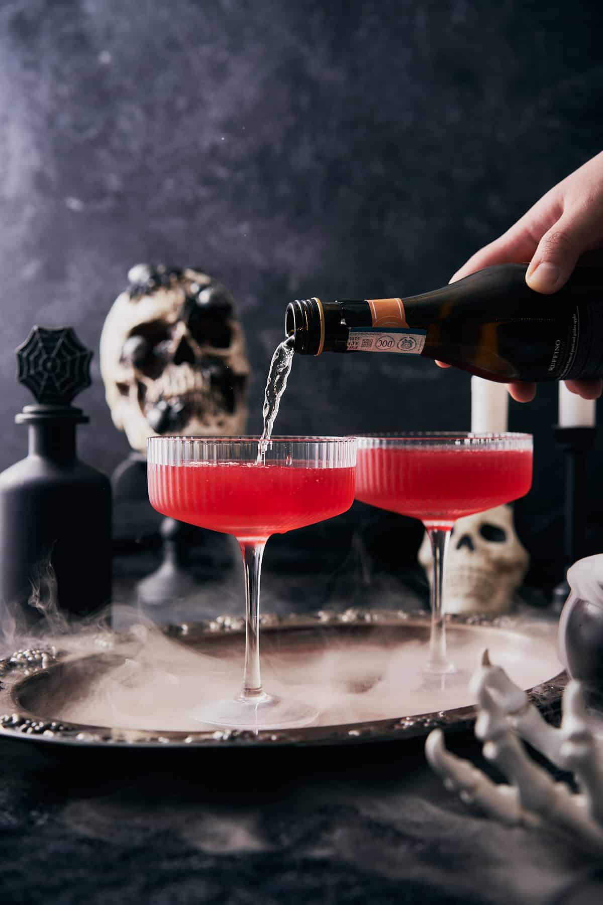 hand pouring sparkling wine into coupe glasses with red liquid inside, on a spooky dark halloween scene with skulls and dry ice.