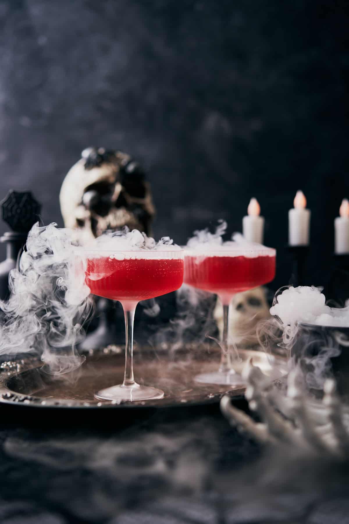 bright red cocktail in a coupe glass with dry ice coming out, with candles, skulls and a skeleton hand in the front of the scene. 