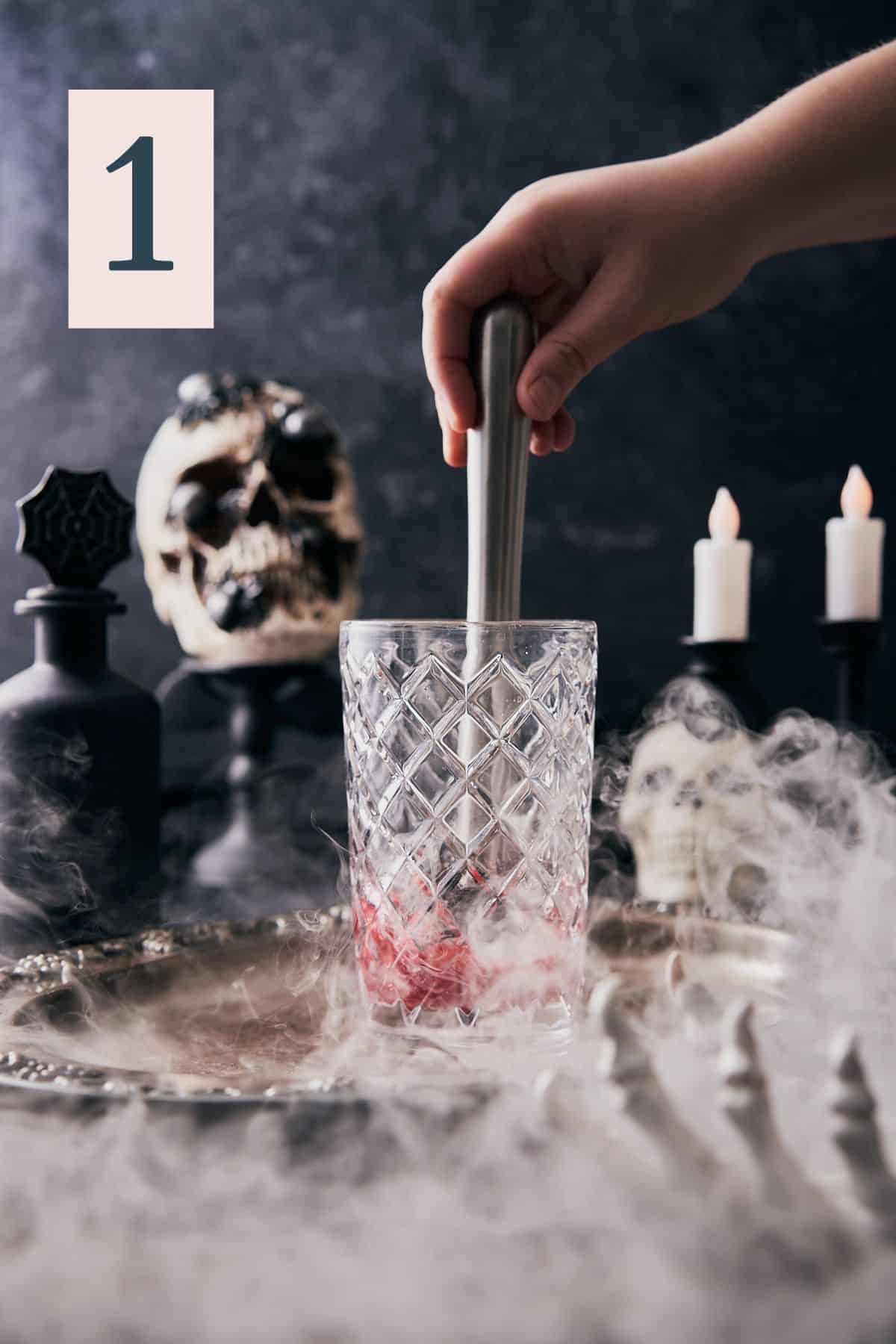 hand muddling raspberries in a glass cocktail shaker, with a very spooky black scene with skulls, candles, and dry ice.