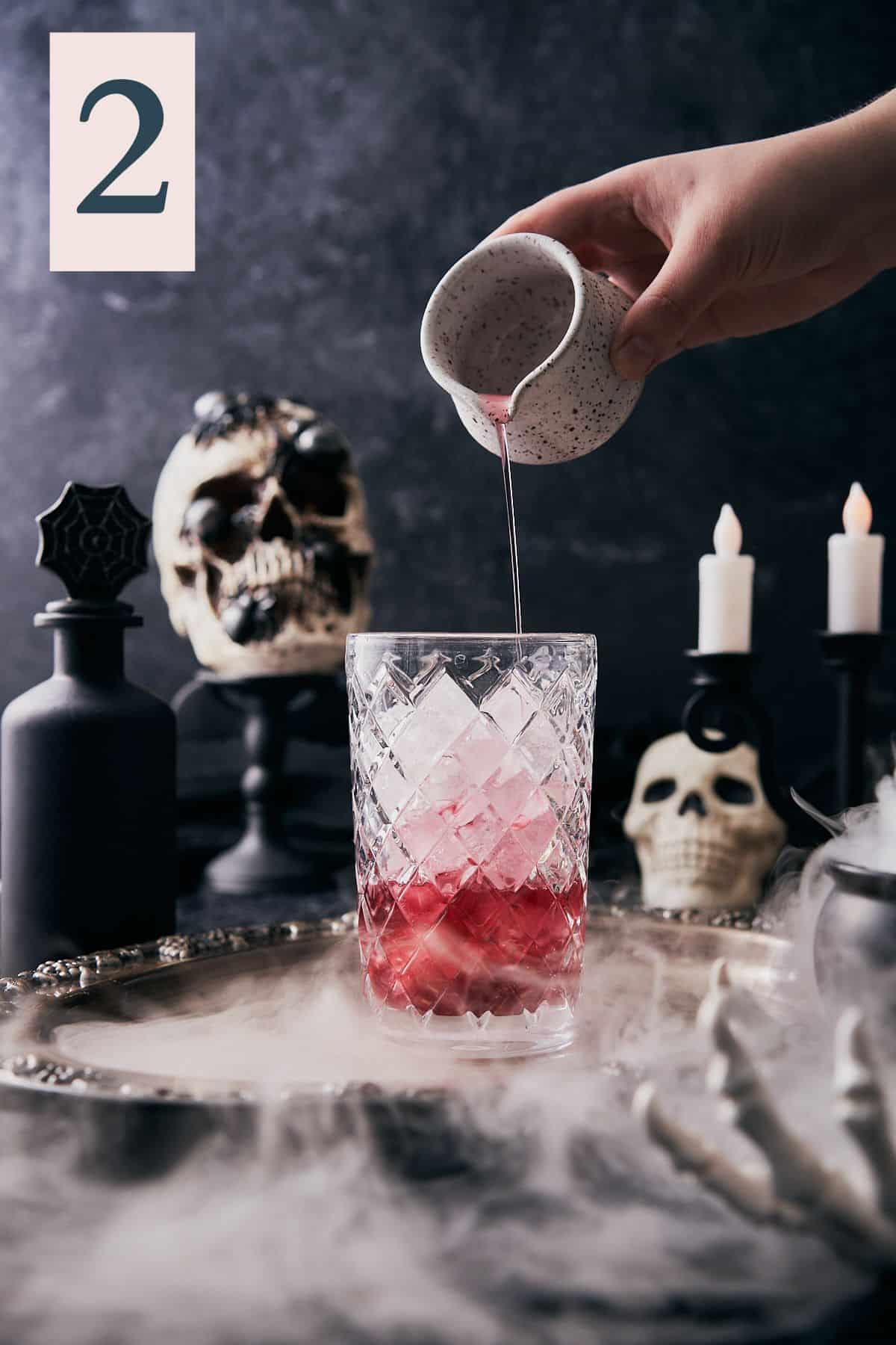 spooky halloween scene with dry ice, and skulls with a hand pouring a red juice into an ice filled cocktail shaker.
